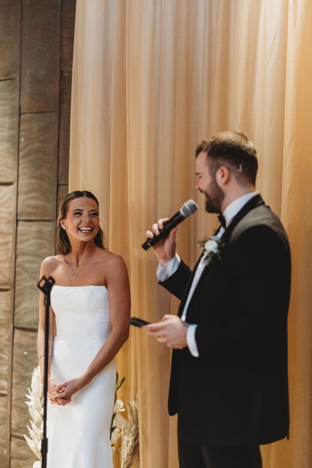 Laughing bride in strapless gown listens as groom speaks into microphone at wedding reception.