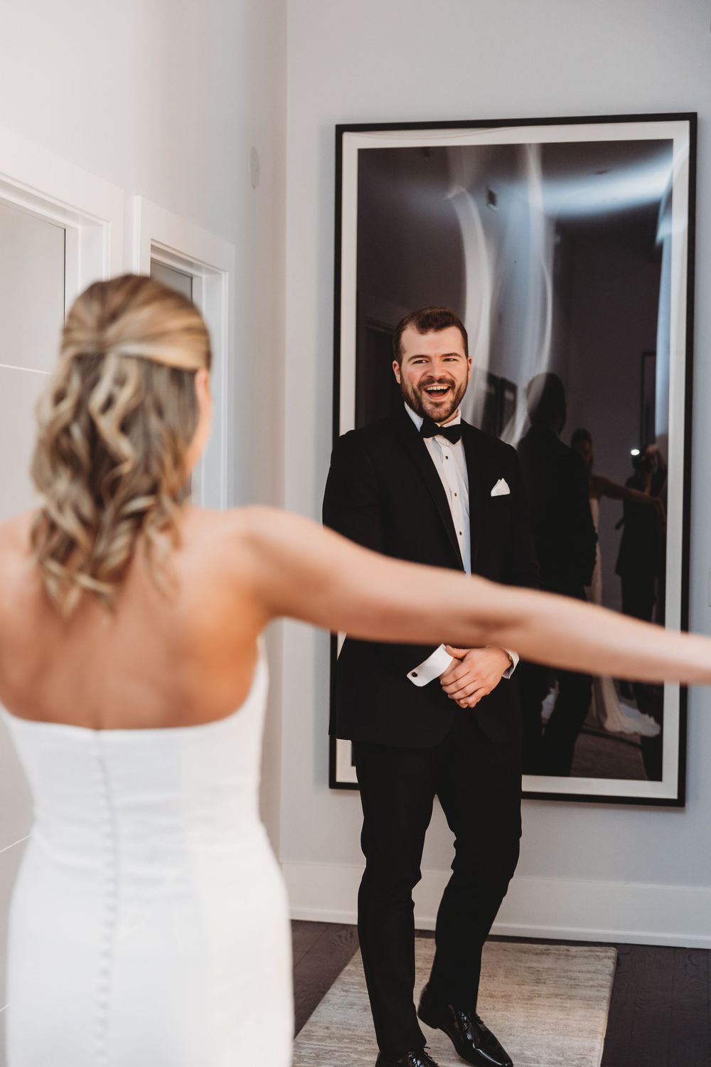 Groom in black tuxedo smiles joyfully as bride approaches him during their wedding day first look reveal.