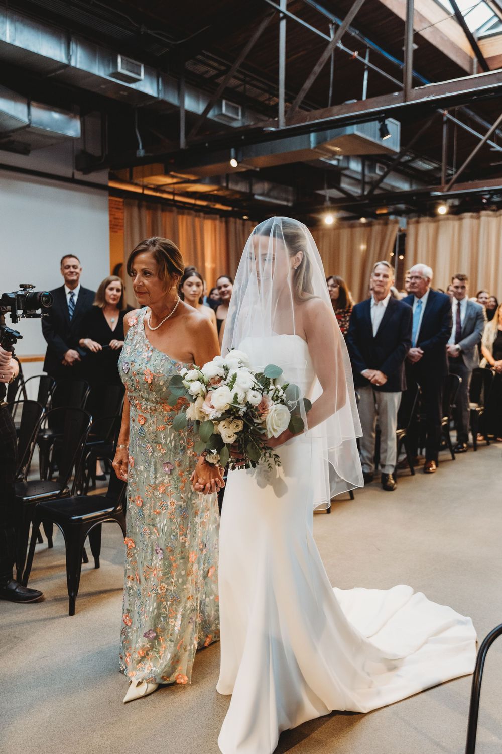 Bride in white gown and veil walks down the aisle with her mother during an indoor wedding ceremony.