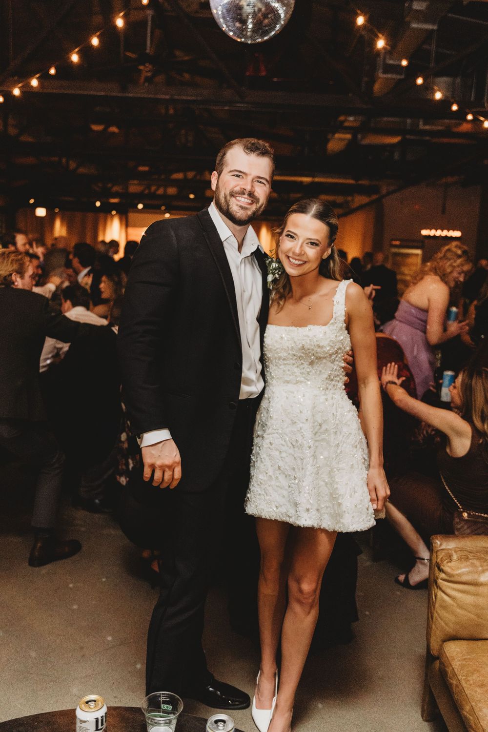 Bride in short white dress and groom in black suit smiling together at indoor wedding reception.