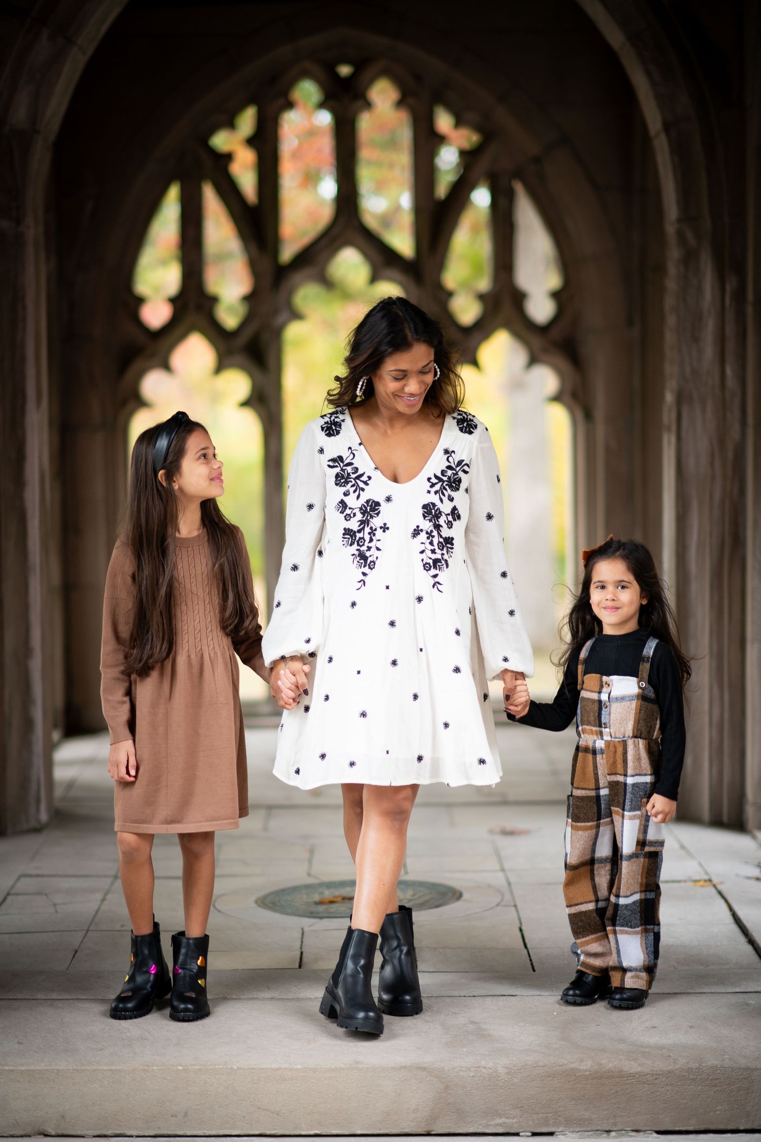 A woman in a white embroidered dress holds hands with two young girls in a gothic archway setting.