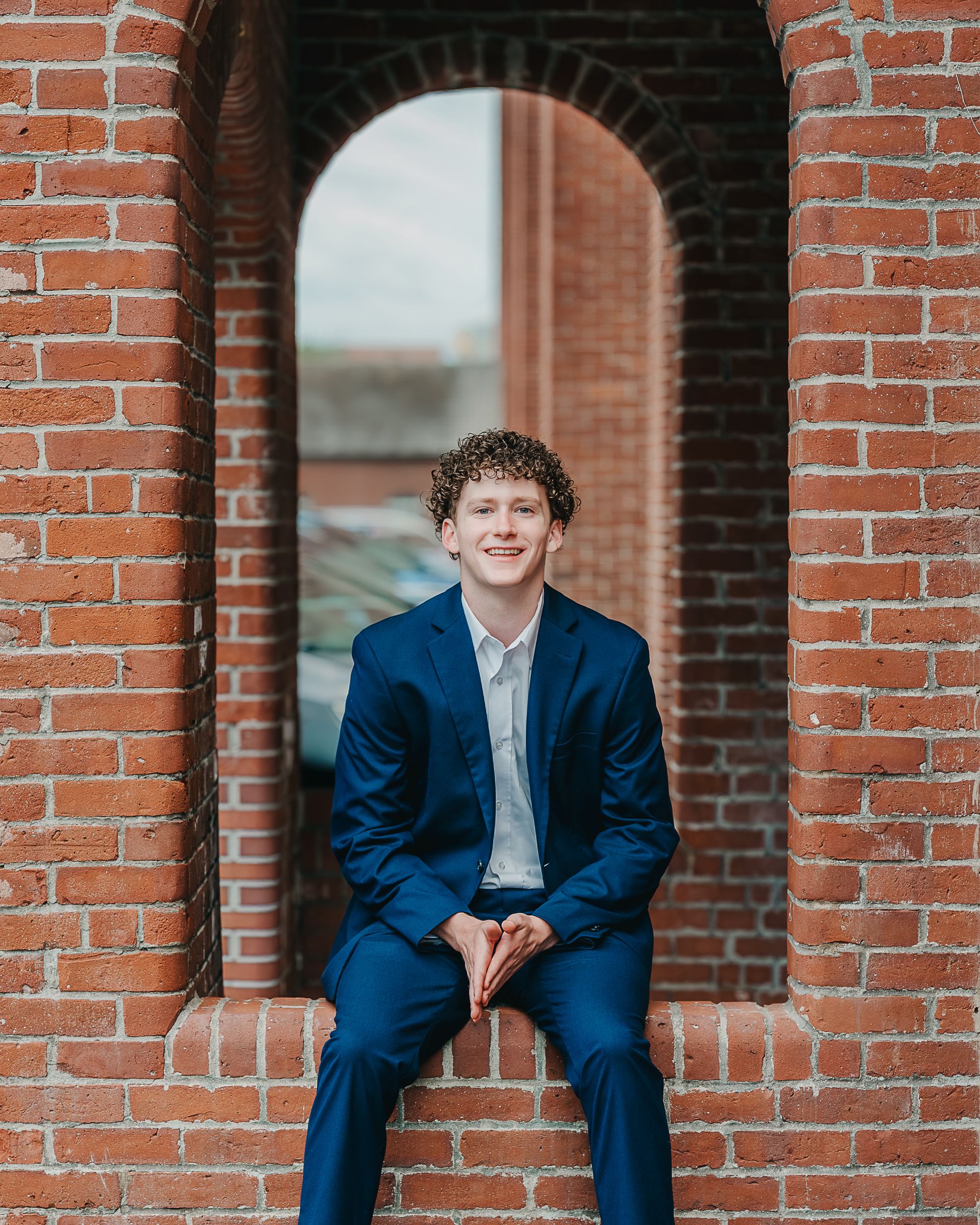 Young man in blue suit sitting in brick archway, smiling confidently for senior portrait photos outdoors.