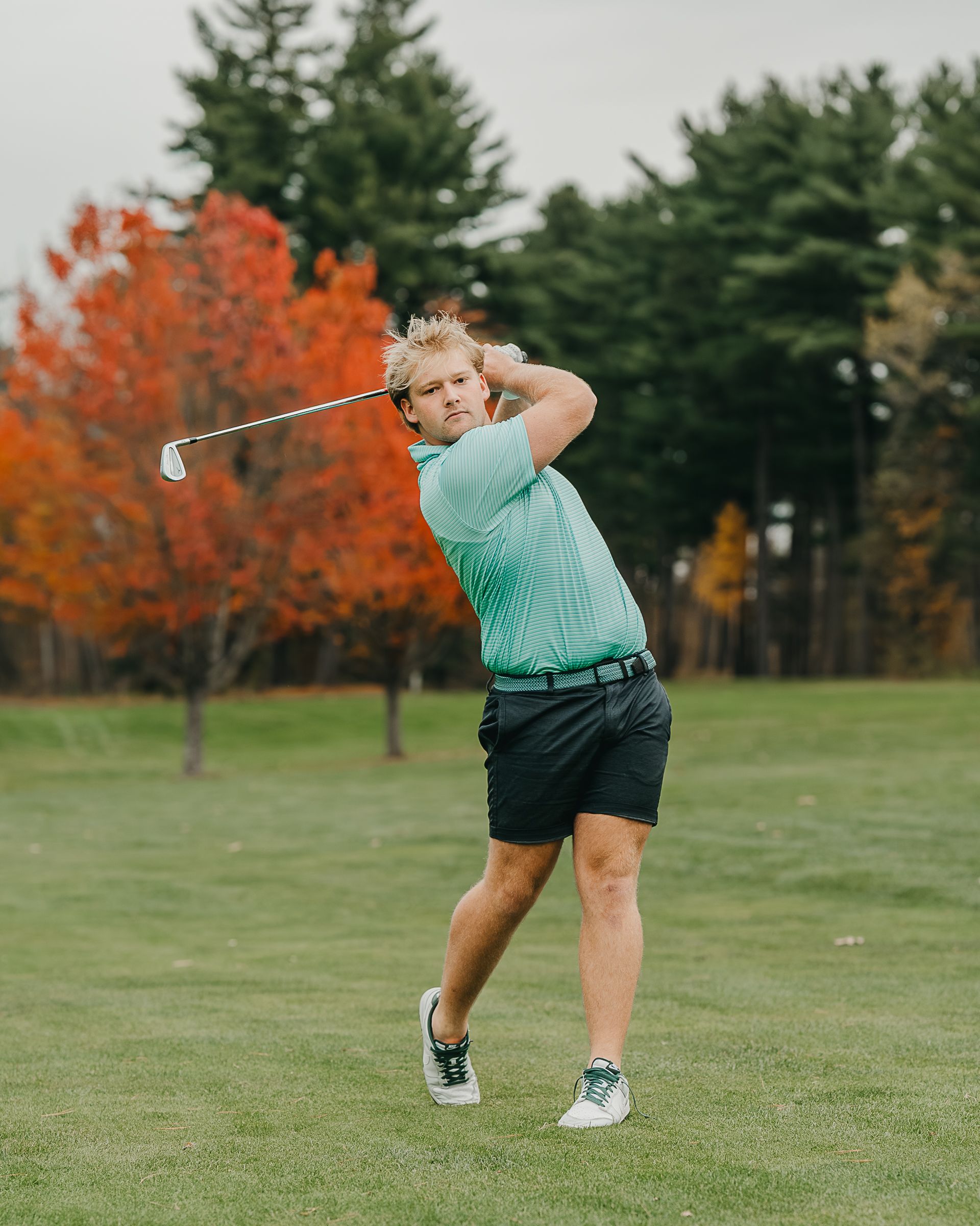 Golfer in mint green shirt swings club on autumn course with colorful red and orange trees in background.