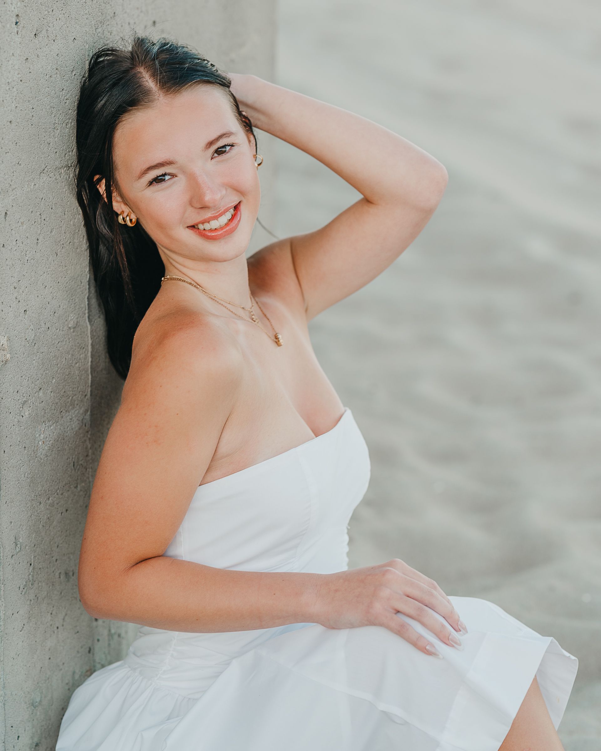 Young woman in white strapless dress smiling outdoors, leaning against concrete wall with hand in dark hair.