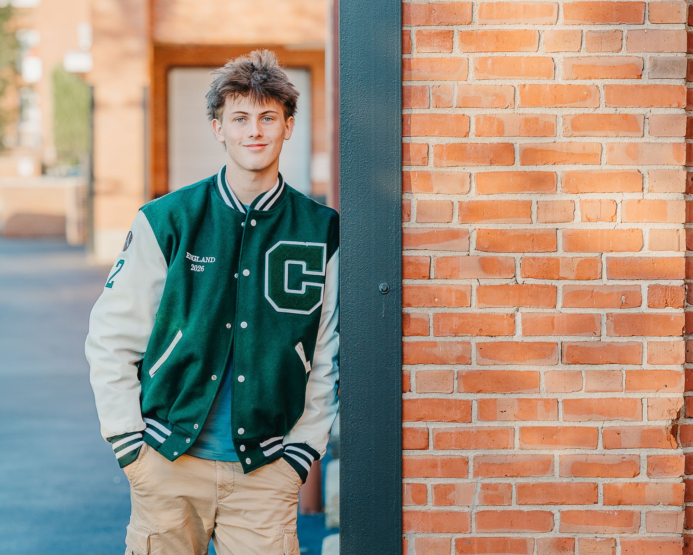 Young man in green varsity jacket posing against a brick wall outdoors.