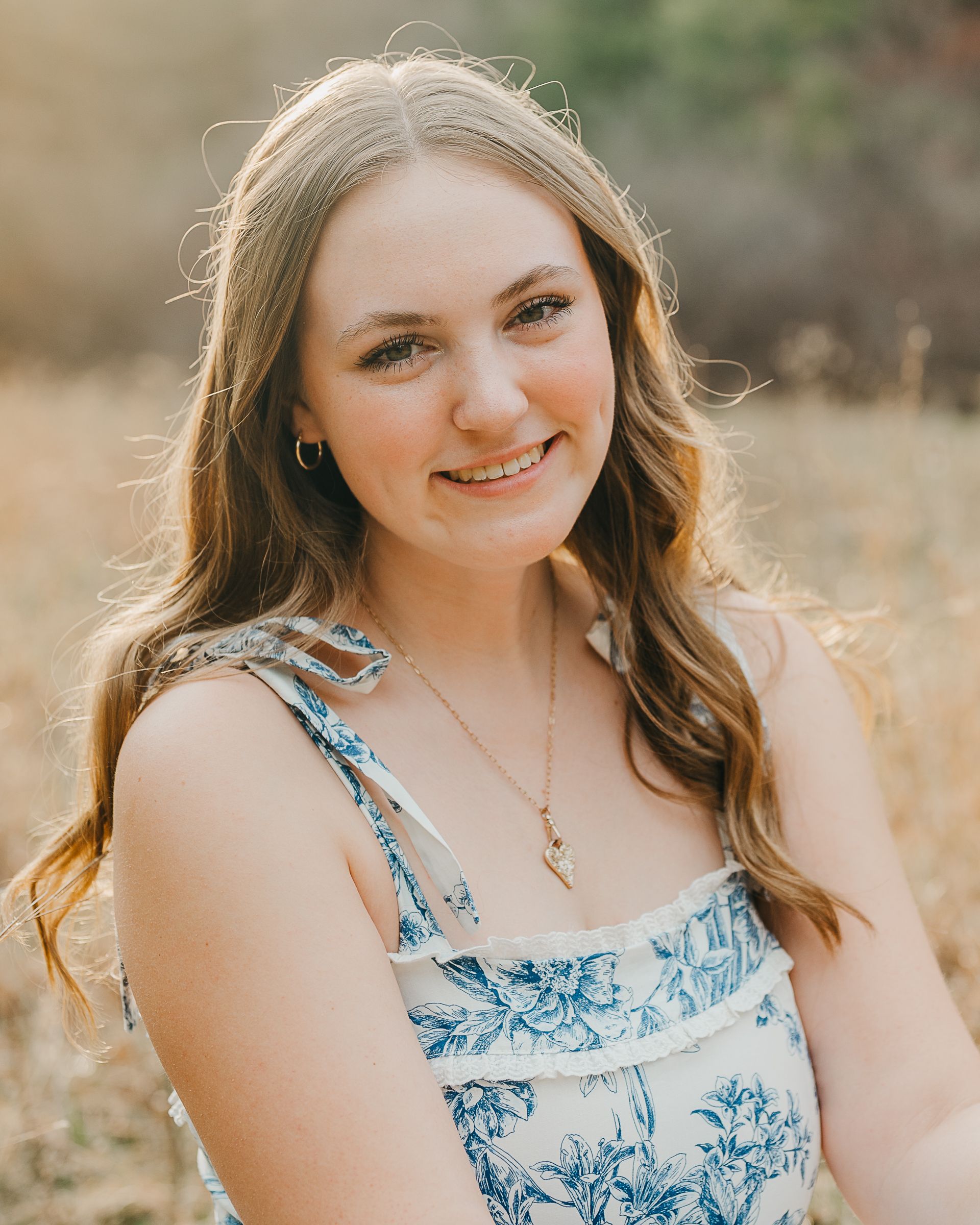 Young woman with long brown hair smiling outdoors, wearing a blue floral dress and gold necklace in a sunlit field.