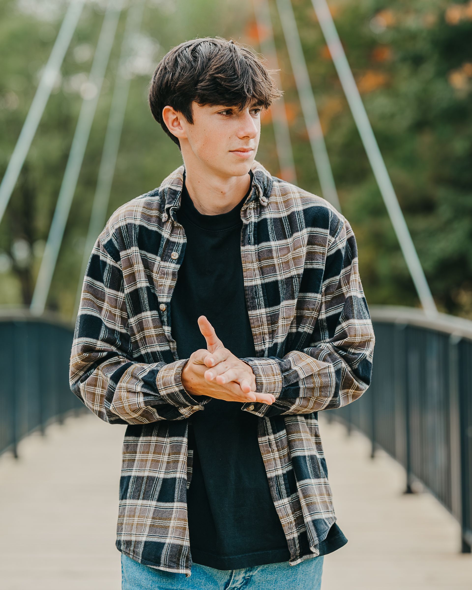 Young man in plaid flannel shirt standing on bridge with arms crossed, looking sideways outdoors.