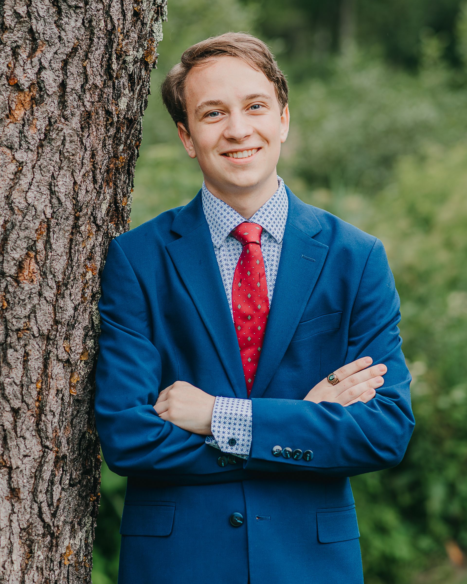 Young man in blue suit and red tie smiling with arms crossed, leaning against a tree outdoors.