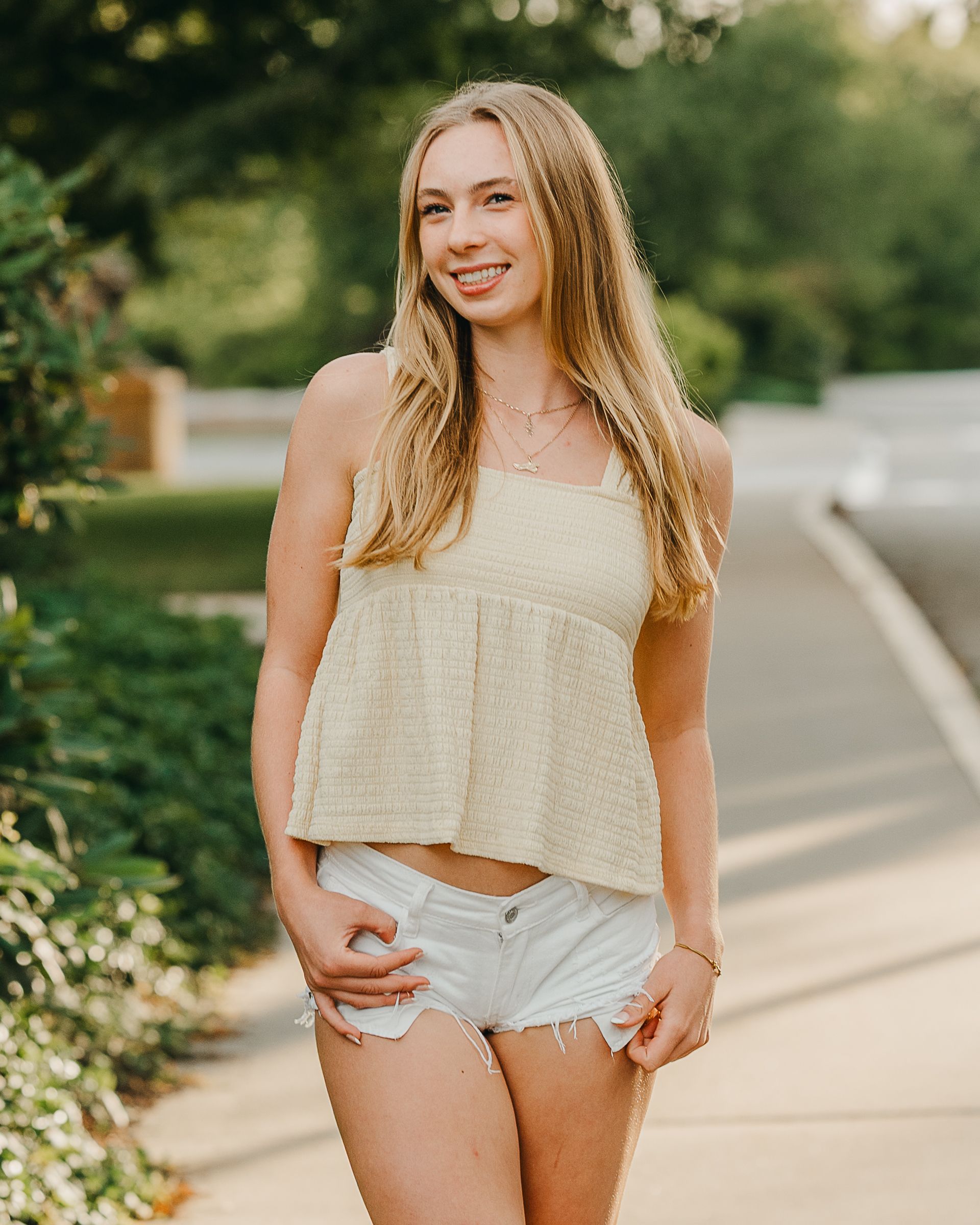Young woman smiling in cream crop top and white denim shorts, posing outdoors on a sunny day.