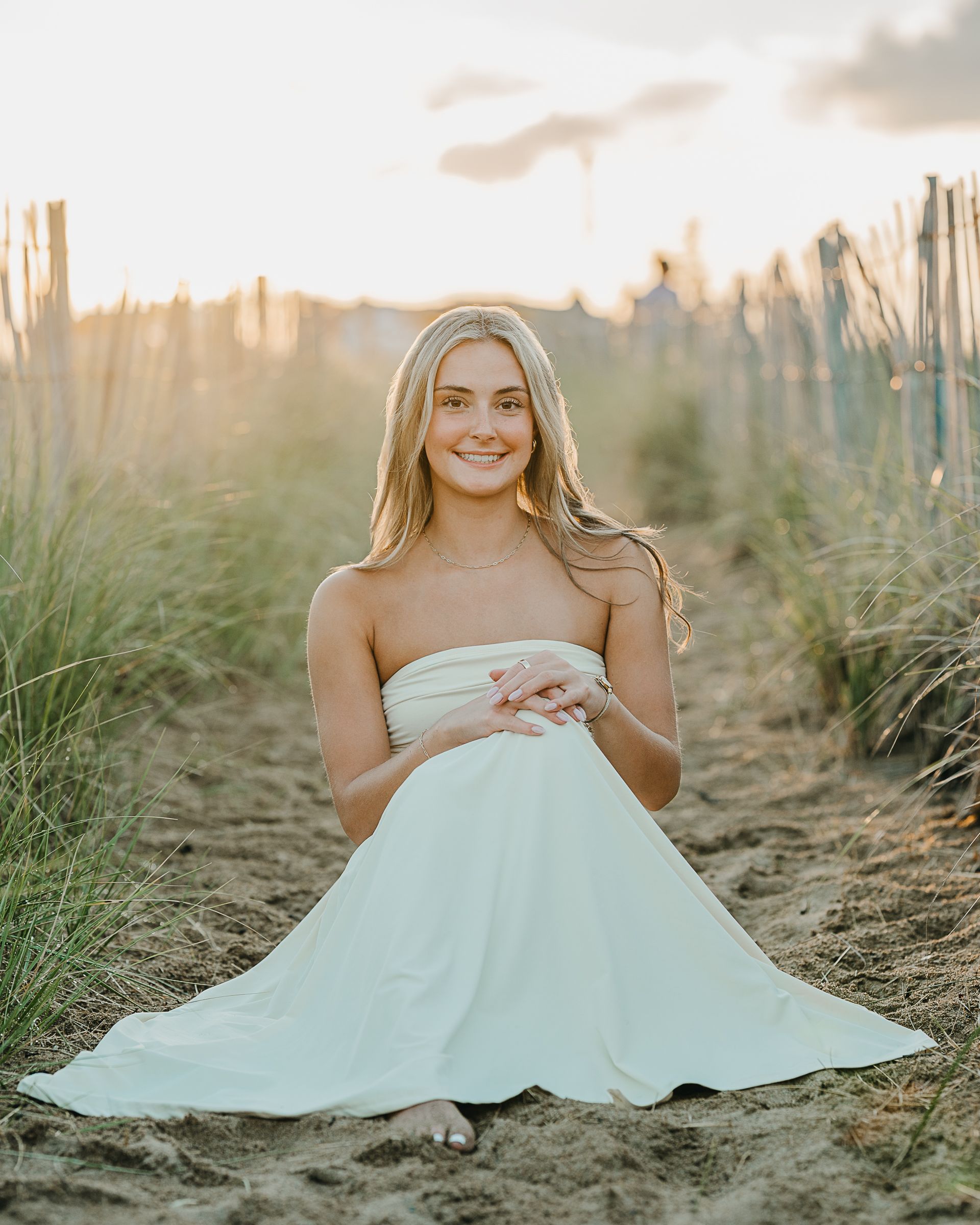 Young blonde woman in white strapless dress sitting on sandy beach path at golden hour sunset.