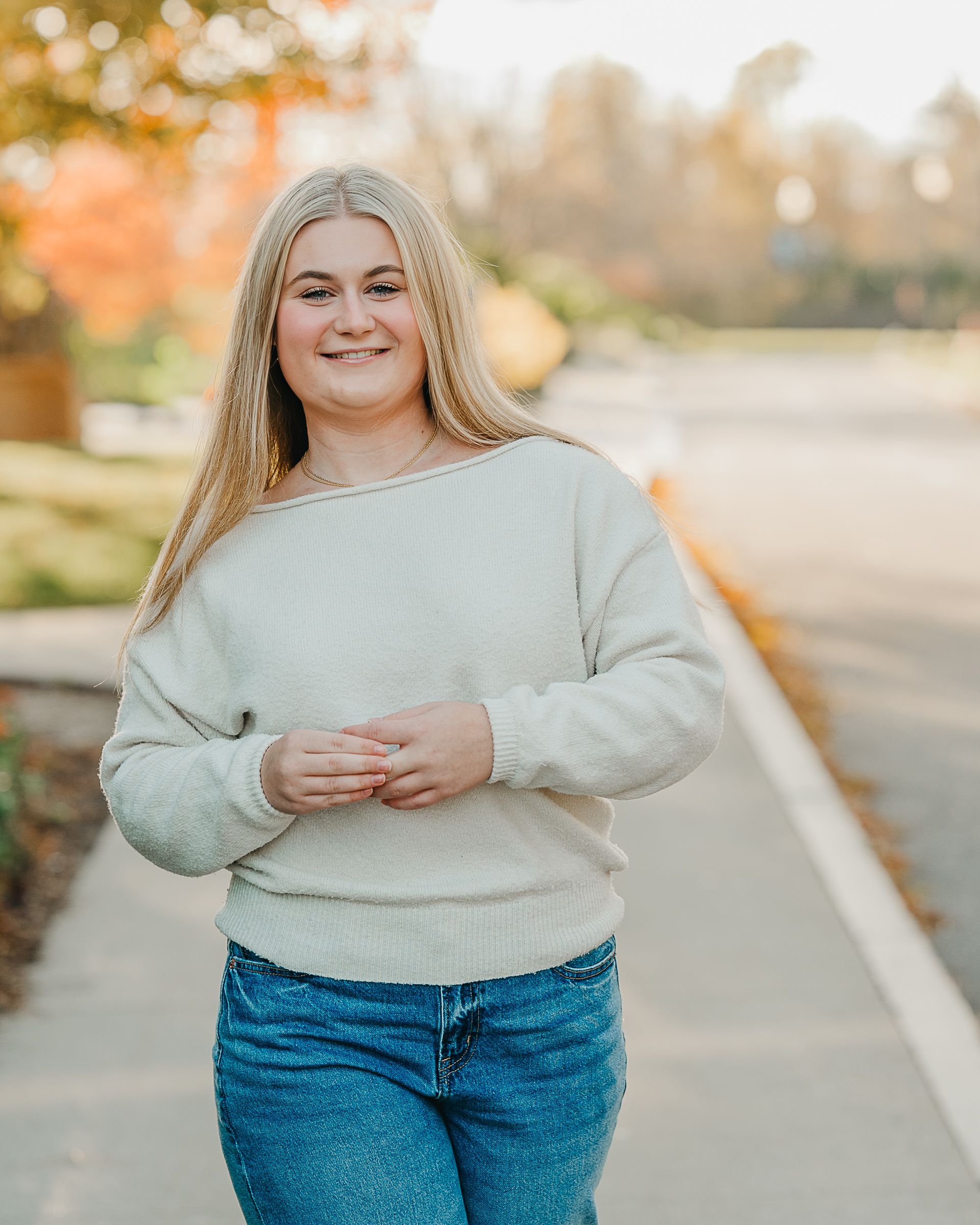 Young blonde woman smiling in a light sage sweatshirt and jeans on a tree-lined path in autumn.