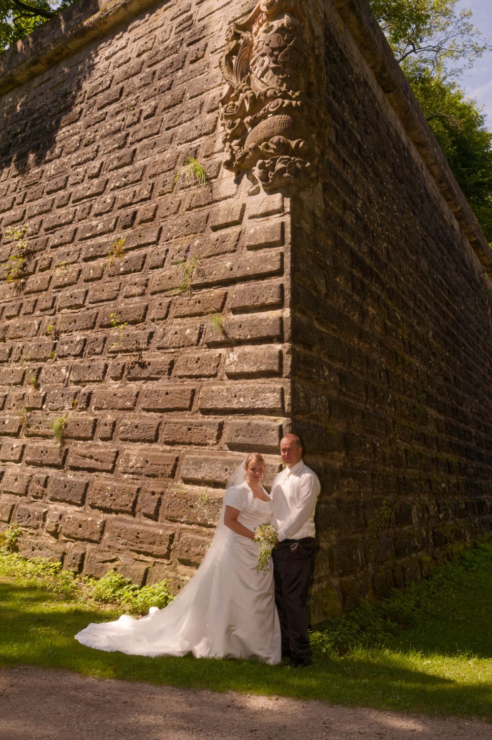 Brautpaar posiert vor einer historischen Steinmauer im Stadtpark Forchheim. Professionelle Hochzeitsfotografie von der Lichtkapsel.