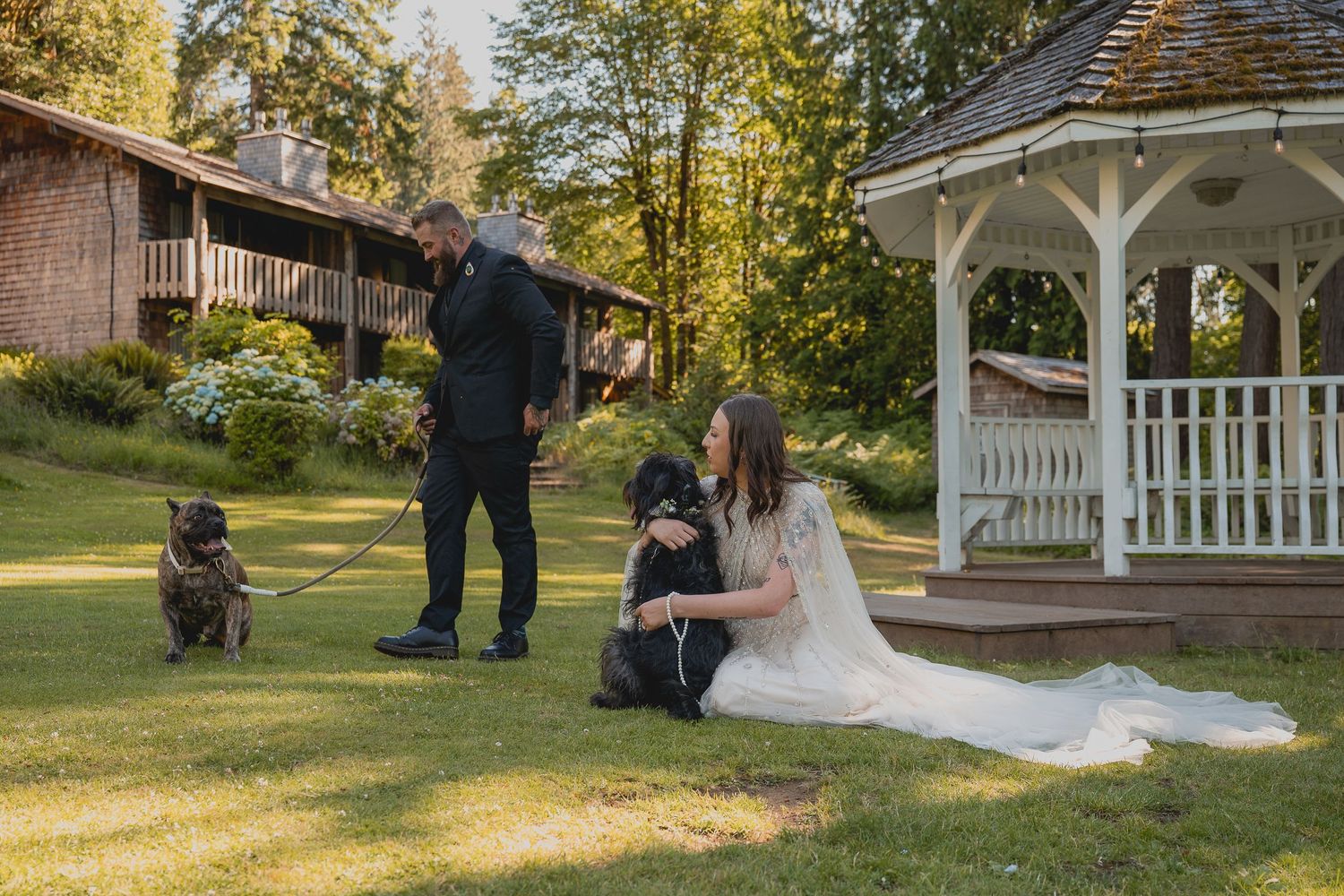 A couple relaxes with their dogs on the lawn in front of a gazebo and rustic lodge.