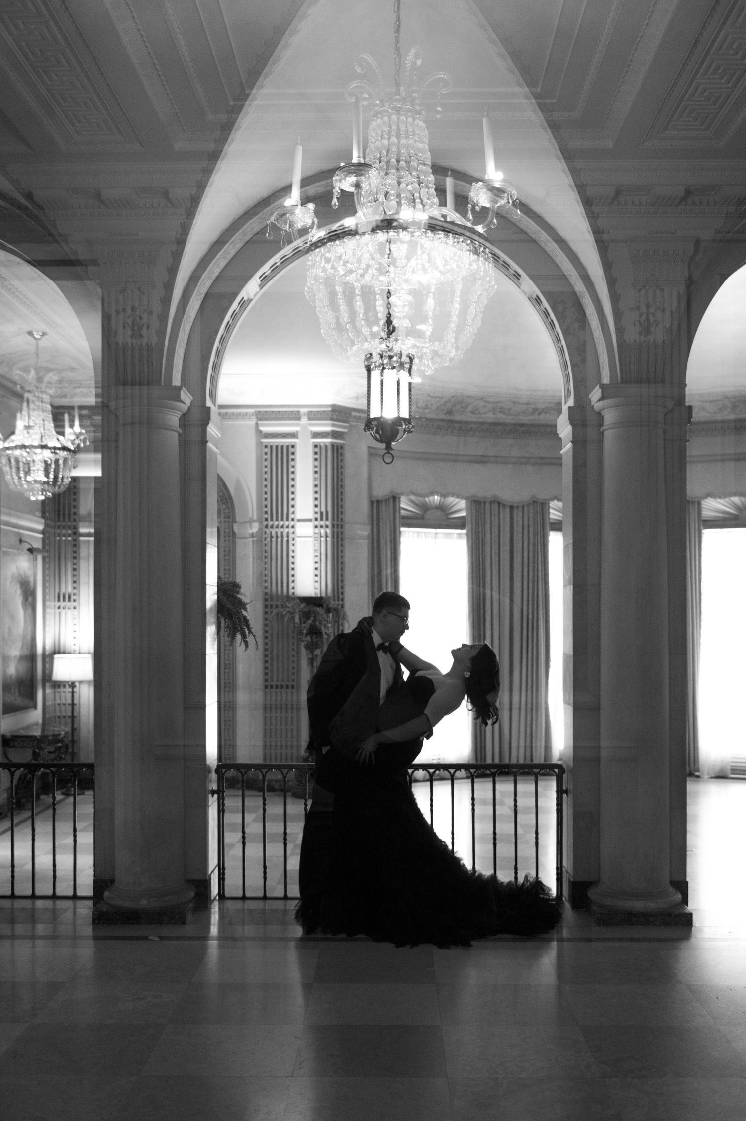 Couple dancing in silhouette under crystal chandelier in elegant ballroom with arched columns, black and white photo.