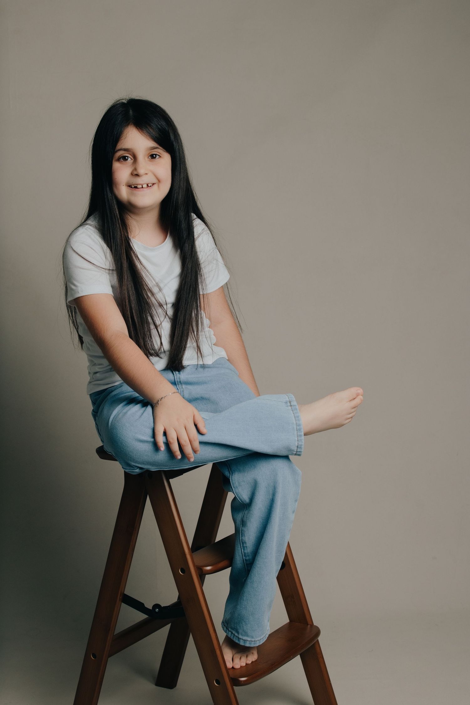Young girl with long dark hair sits on wooden stool, wearing white tee and blue jeans, smiling at camera.