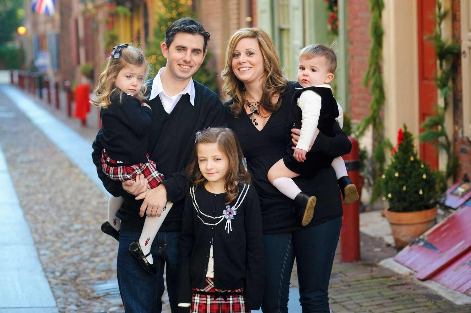 Family of five posing on a charming brick street during Christmas, parents holding toddlers with young girl in center.