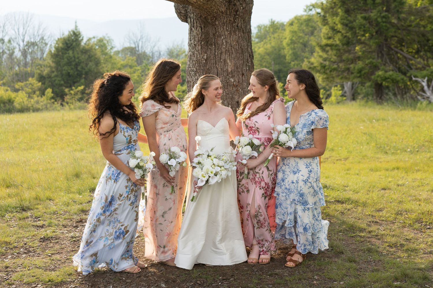 Bride in white gown with four bridesmaids in floral dresses holding bouquets, standing by a tree in a green field.