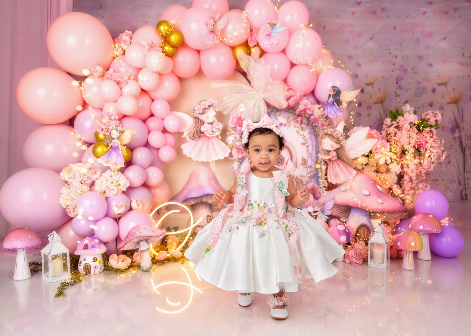 Baby girl in white floral dress stands before pink balloon arch with mushroom decorations at first birthday party.