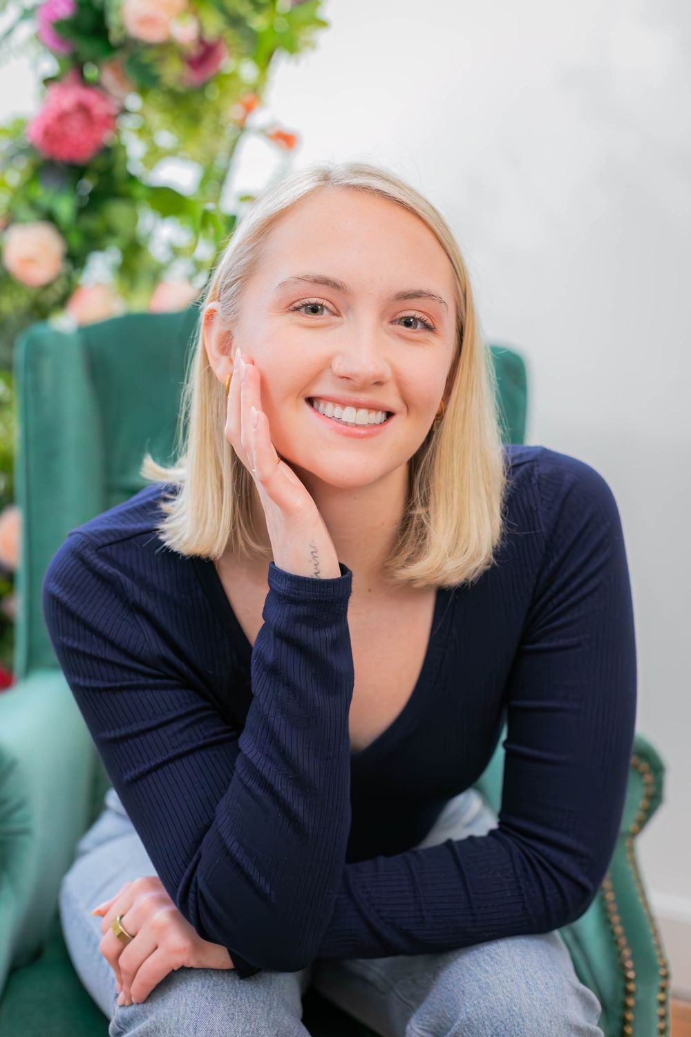 A woman in a navy sweater sits in a green armchair with floral decorations in the background.