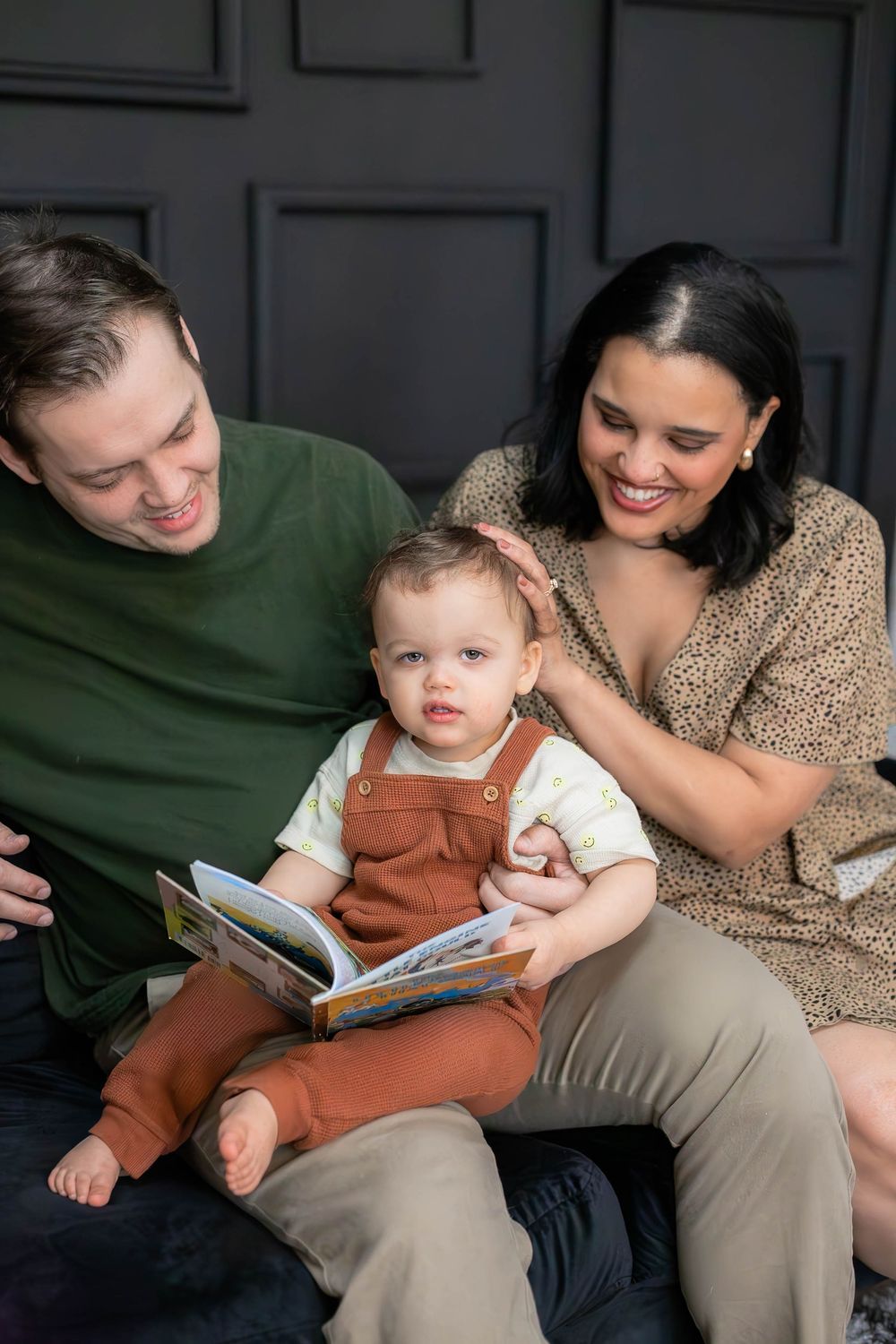Family enjoying storytime together on a couch with a small child in brown overalls reading a book.