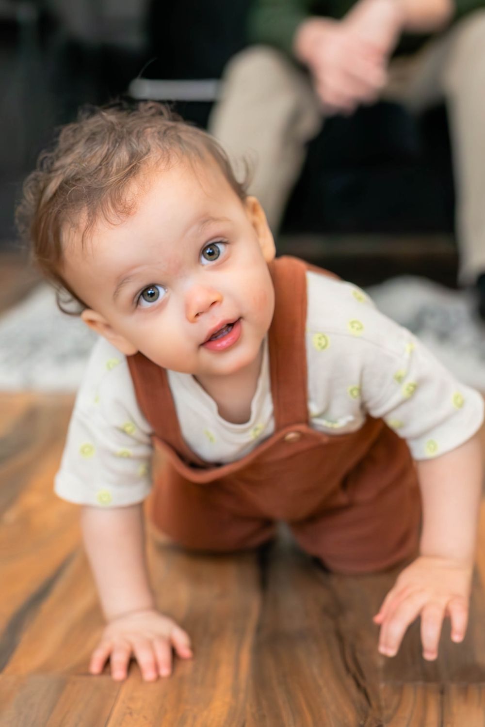 Sweet toddler in brown overalls and floral shirt crawls on hardwood floor with eager expression.