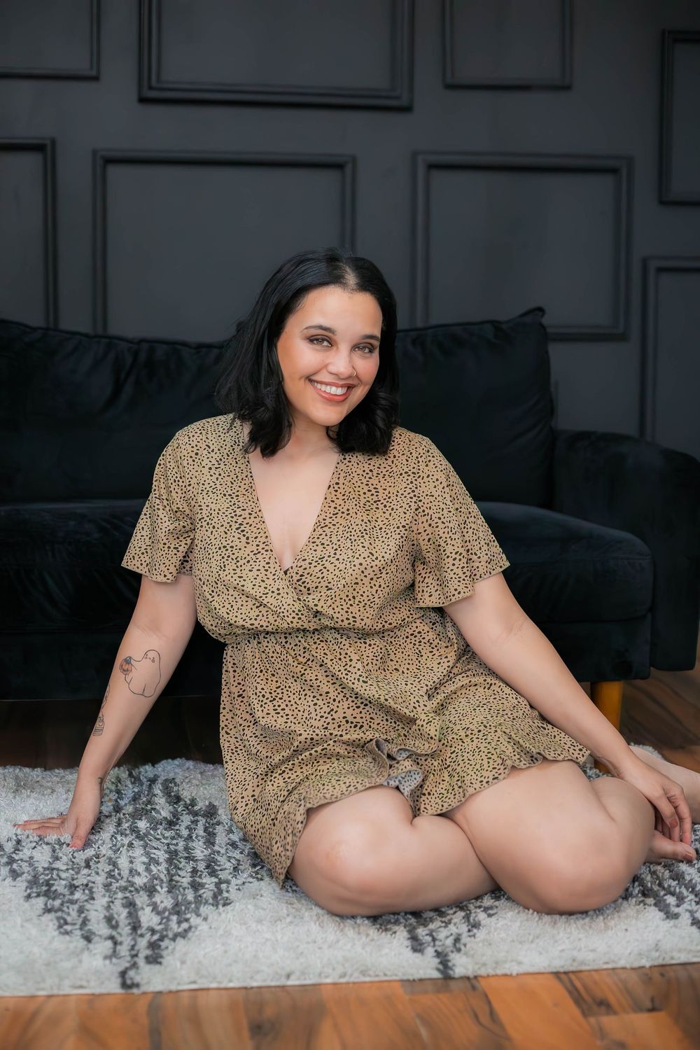 Smiling woman in leopard print dress sits on plush grey rug against dark wall with decorative molding.