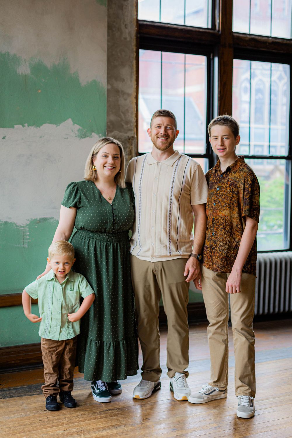 A family of four poses in coordinating vintage-inspired outfits in front of a large window with green painted wall.