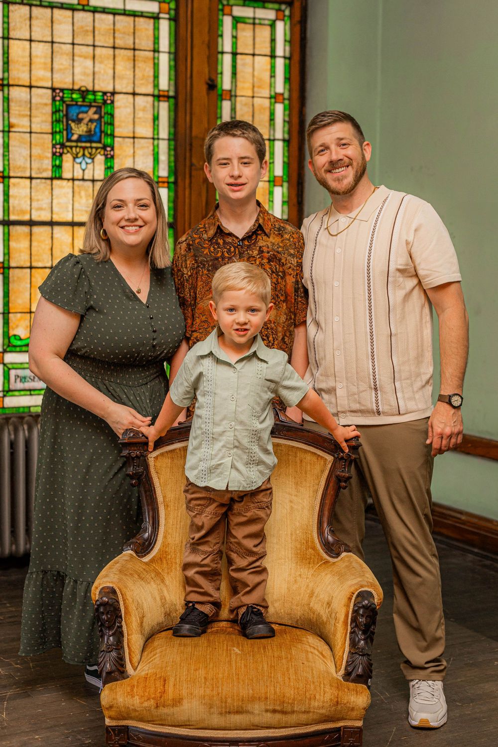 A family poses together in front of a stained glass window with a vintage gold chair in an interior setting.