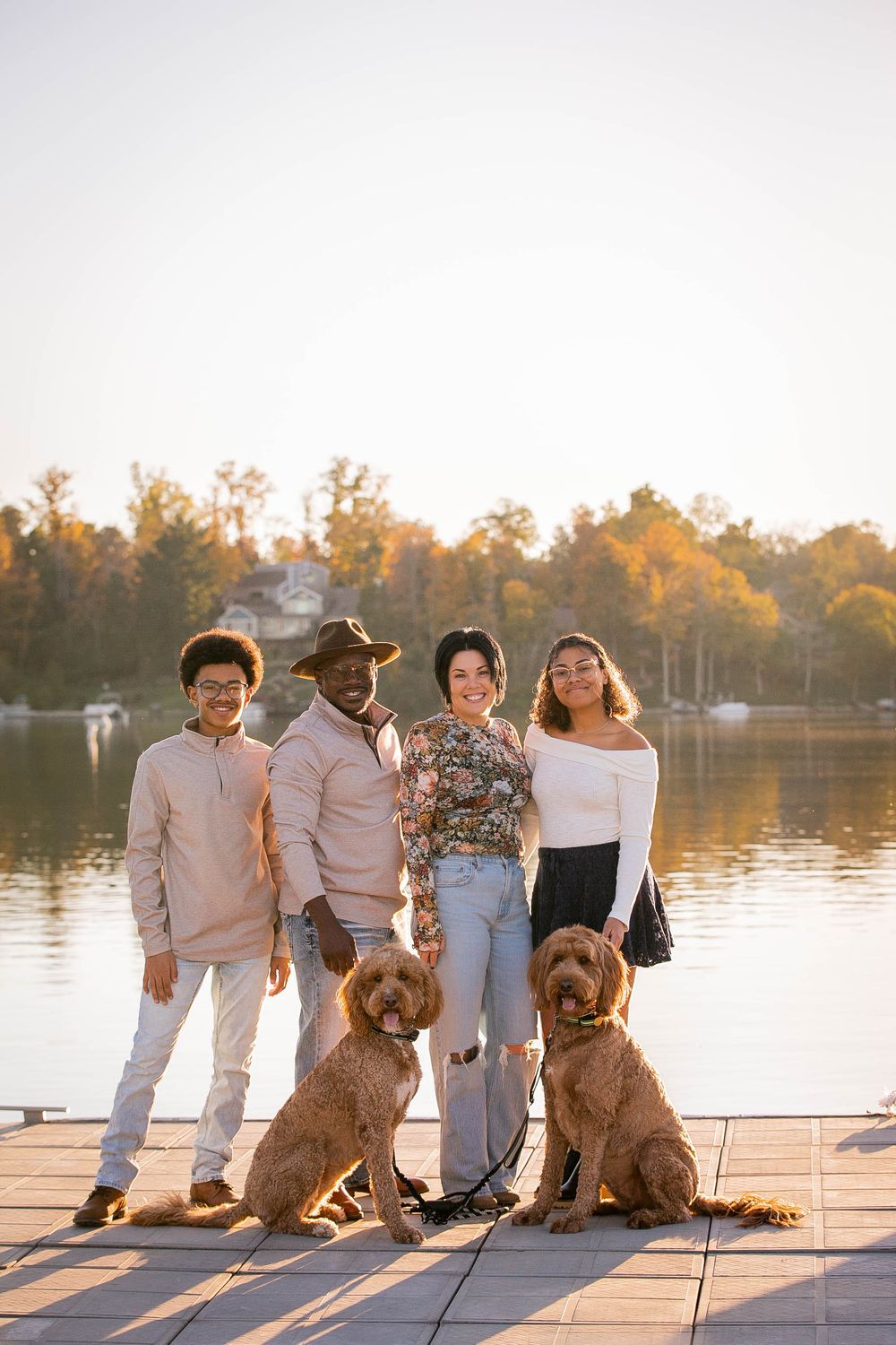 A group in casual attire poses on a lakeside dock with two golden-colored dogs at sunset.