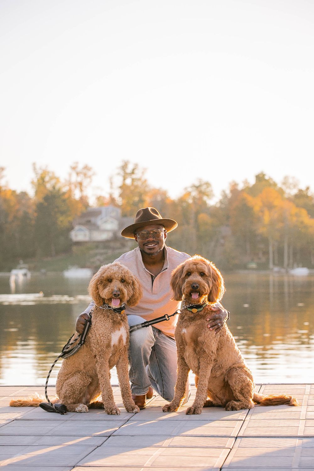 Two golden doodle dogs sit obediently with their owner on a lakeside dock at sunset.