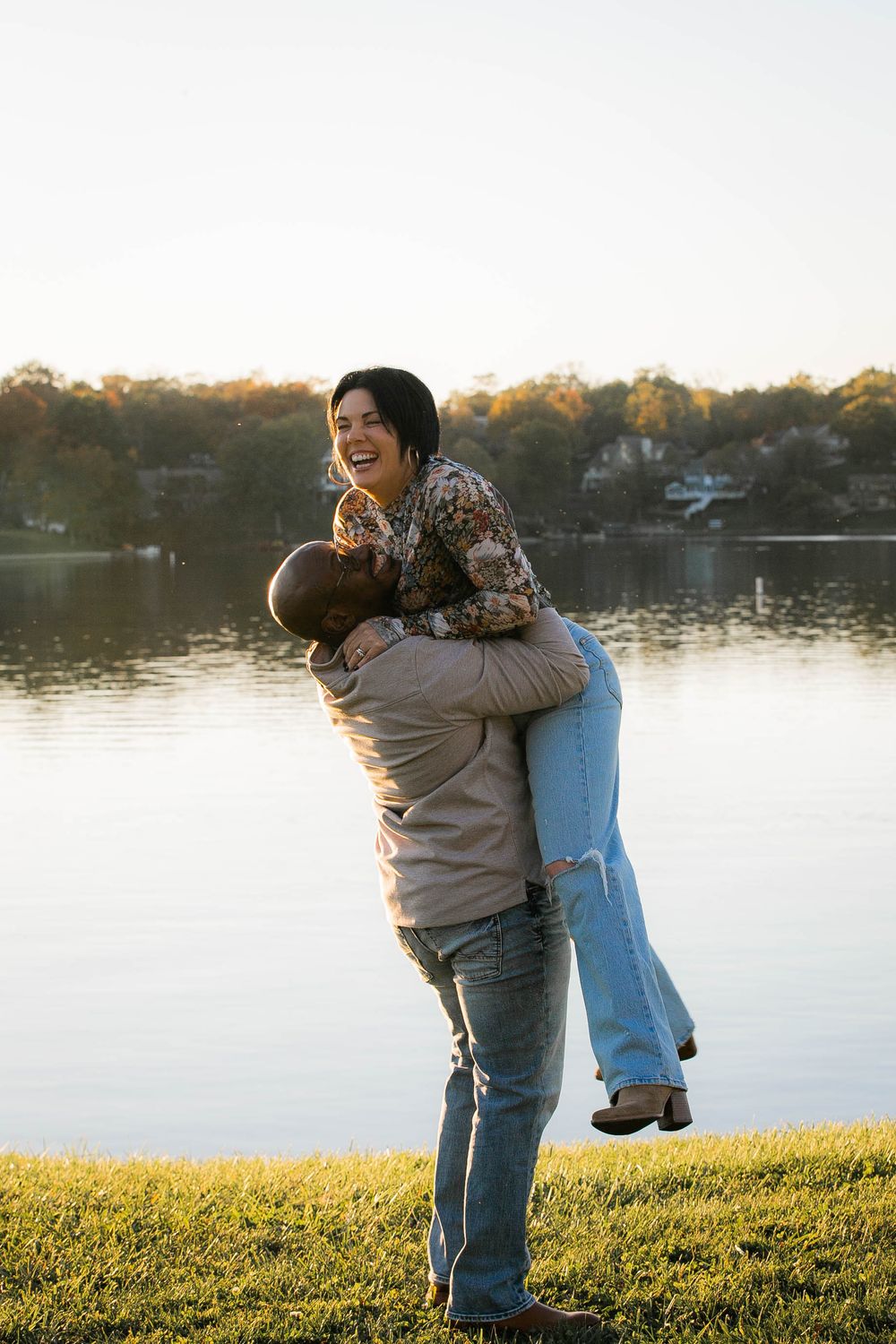 A joyful embrace between two people by a serene lake at sunset on a grassy shoreline.