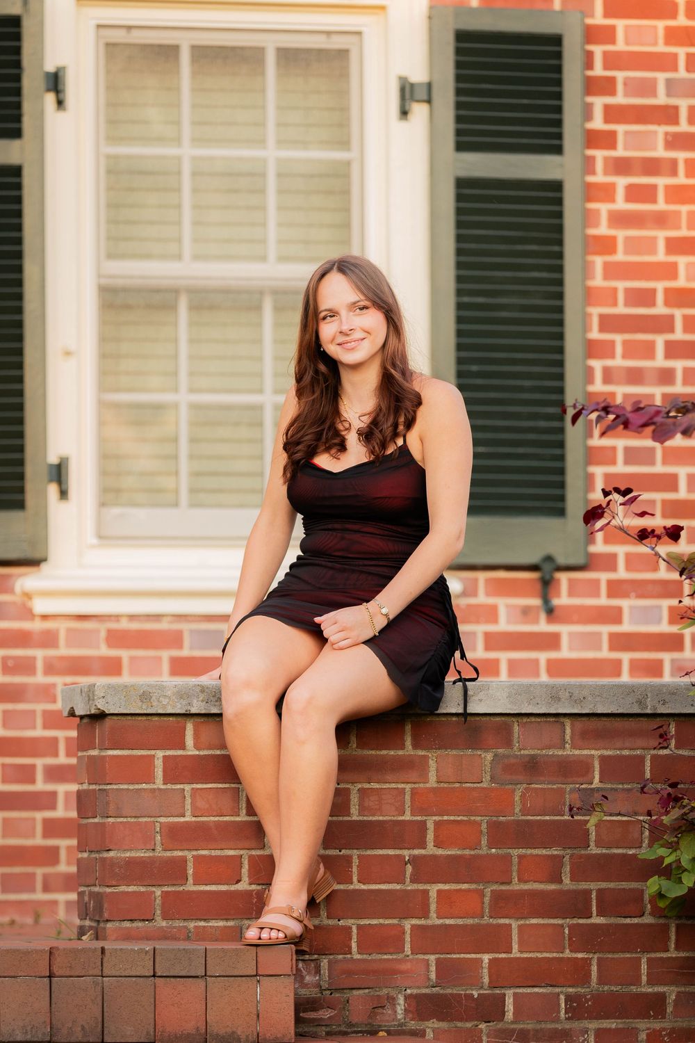 A woman in a black dress sits on a brick ledge in front of a traditional brick house with green shutters.
