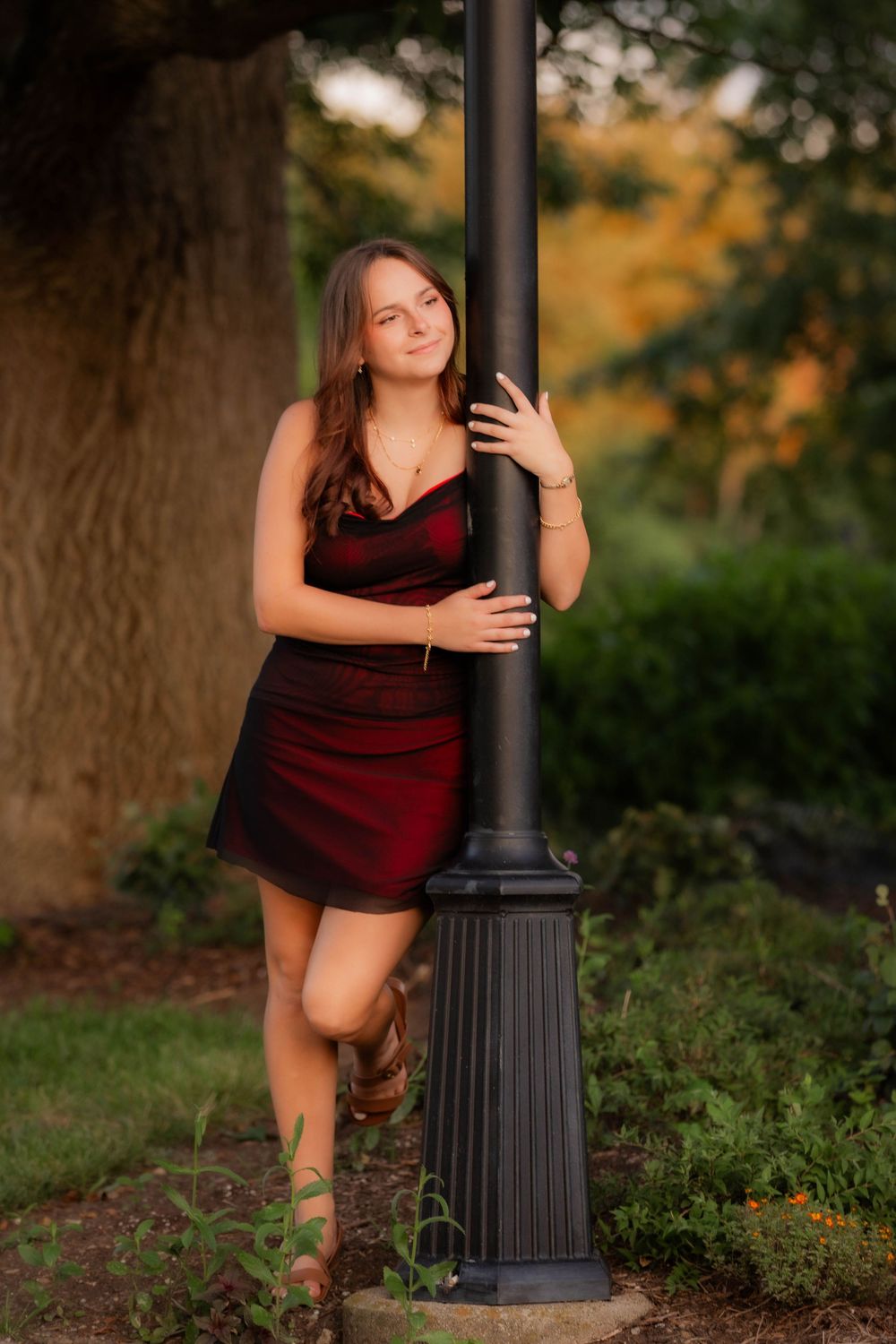 A figure in a burgundy dress poses gracefully against a black lamppost in a garden setting during golden hour.