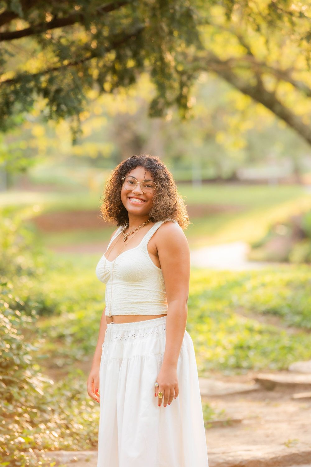A person in a flowing white dress poses gracefully outdoors in golden sunlit garden scenery.