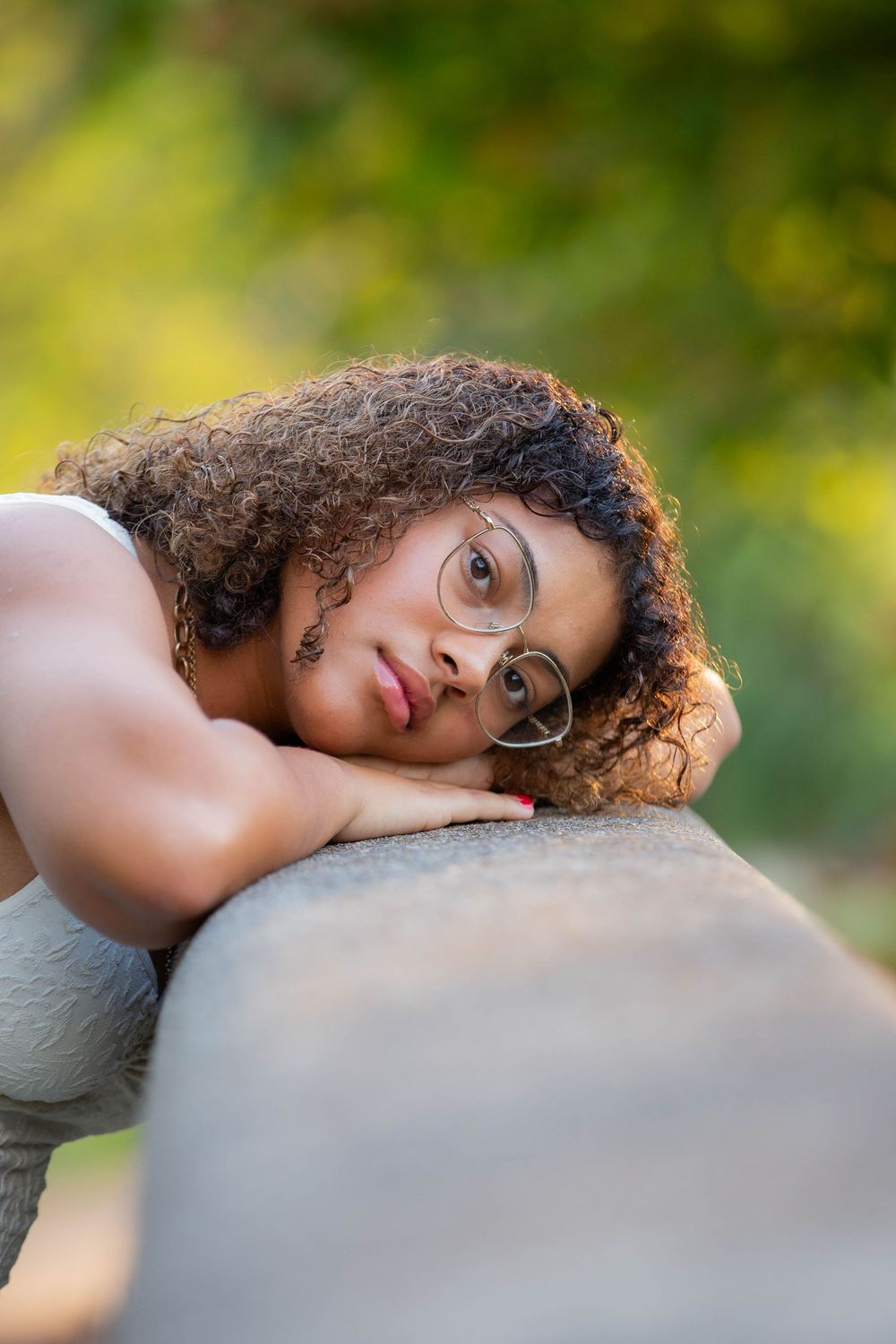 A person with curly hair and glasses rests their head against a light gray surface in warm natural lighting.