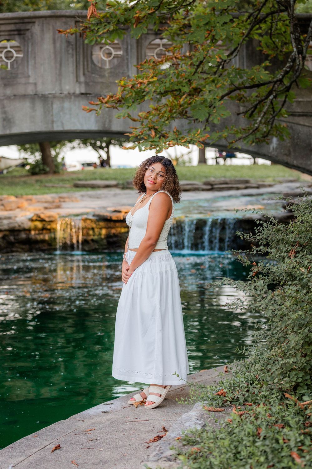 A white sundress-clad figure stands near a tranquil pool with stone architecture and greenery in the background.