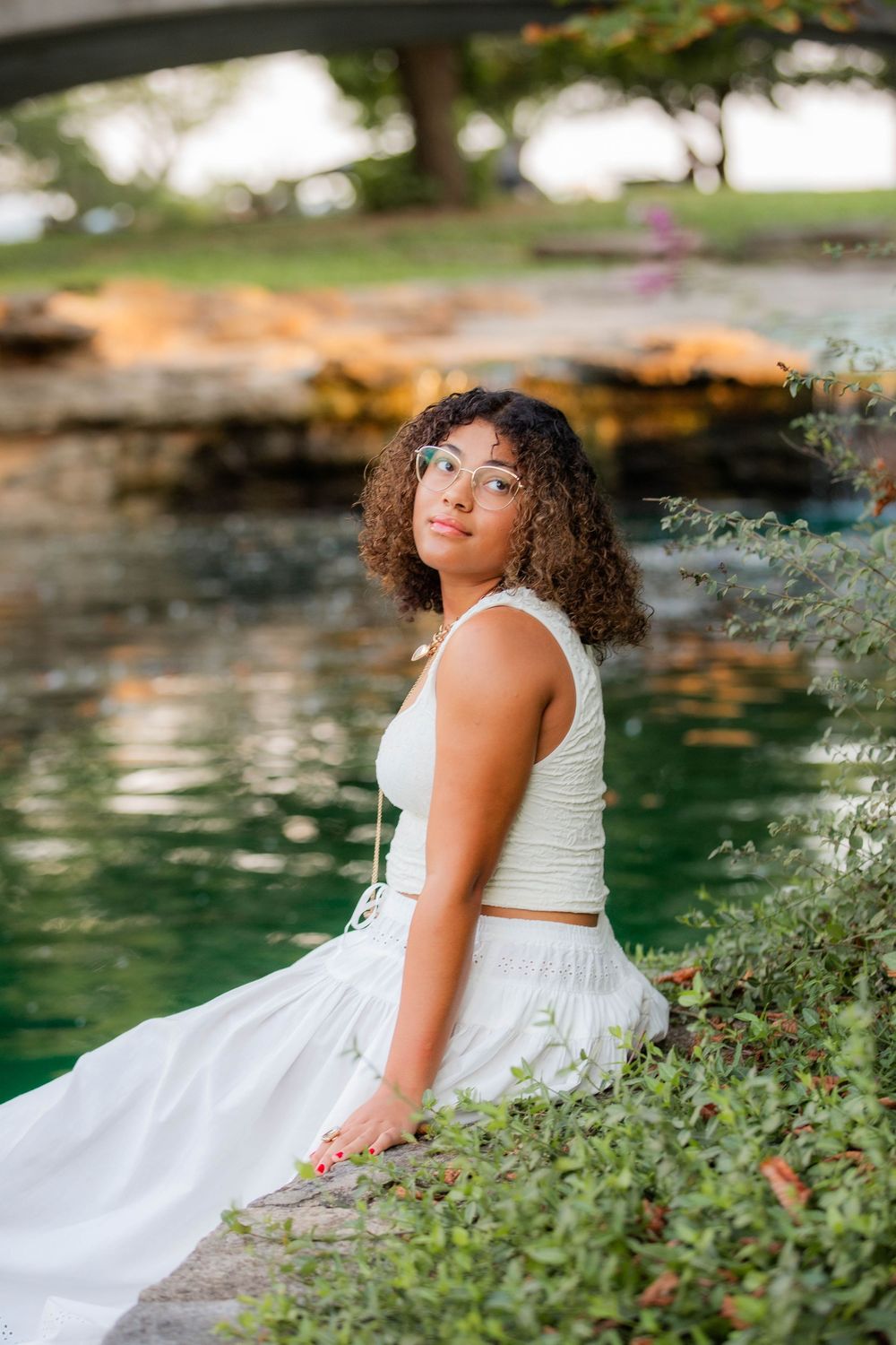 A white dress flows elegantly against lush greenery and a peaceful pond in a serene garden setting.