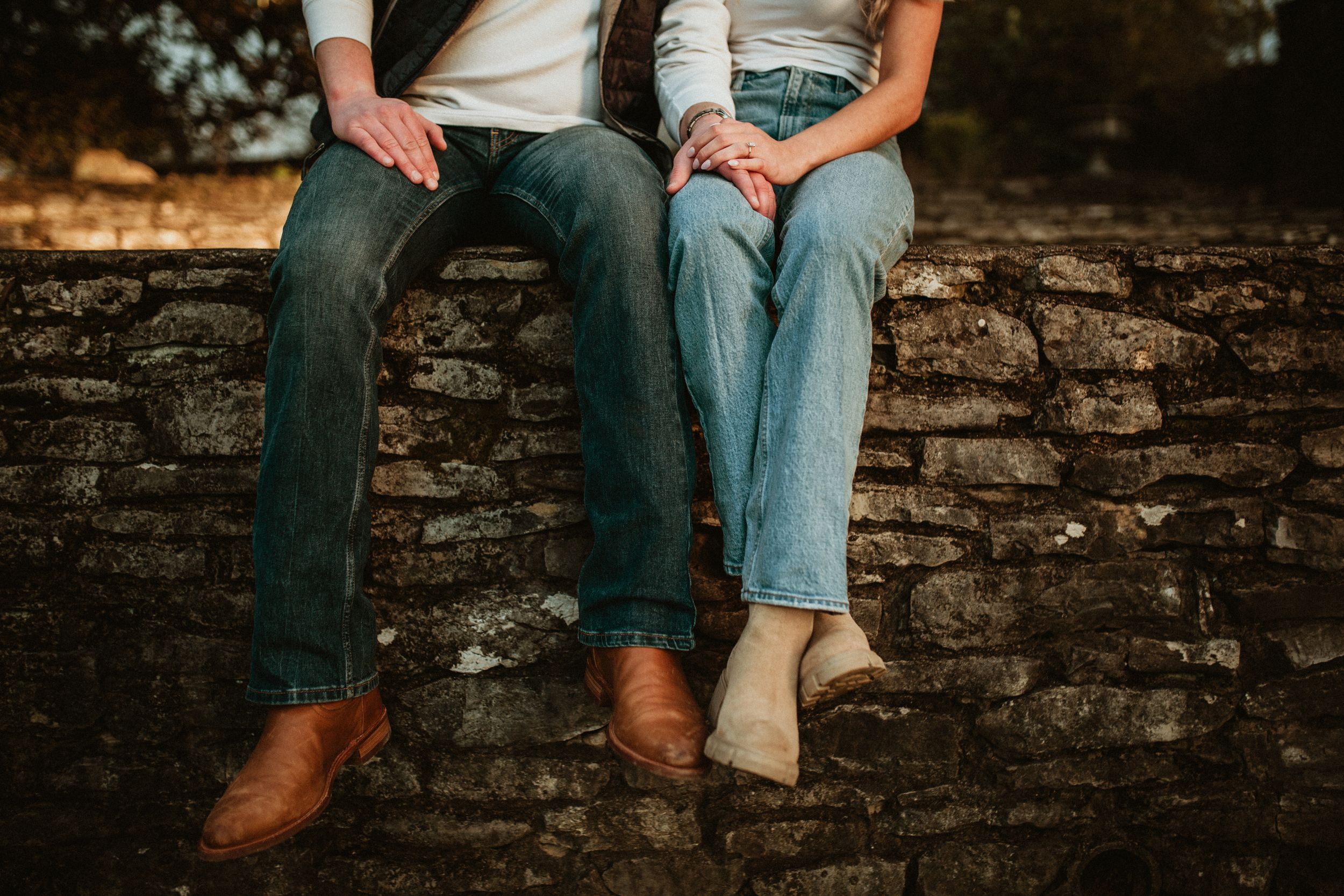 A couple sits casually on a rustic brick wall wearing jeans and brown boots in warm lighting.