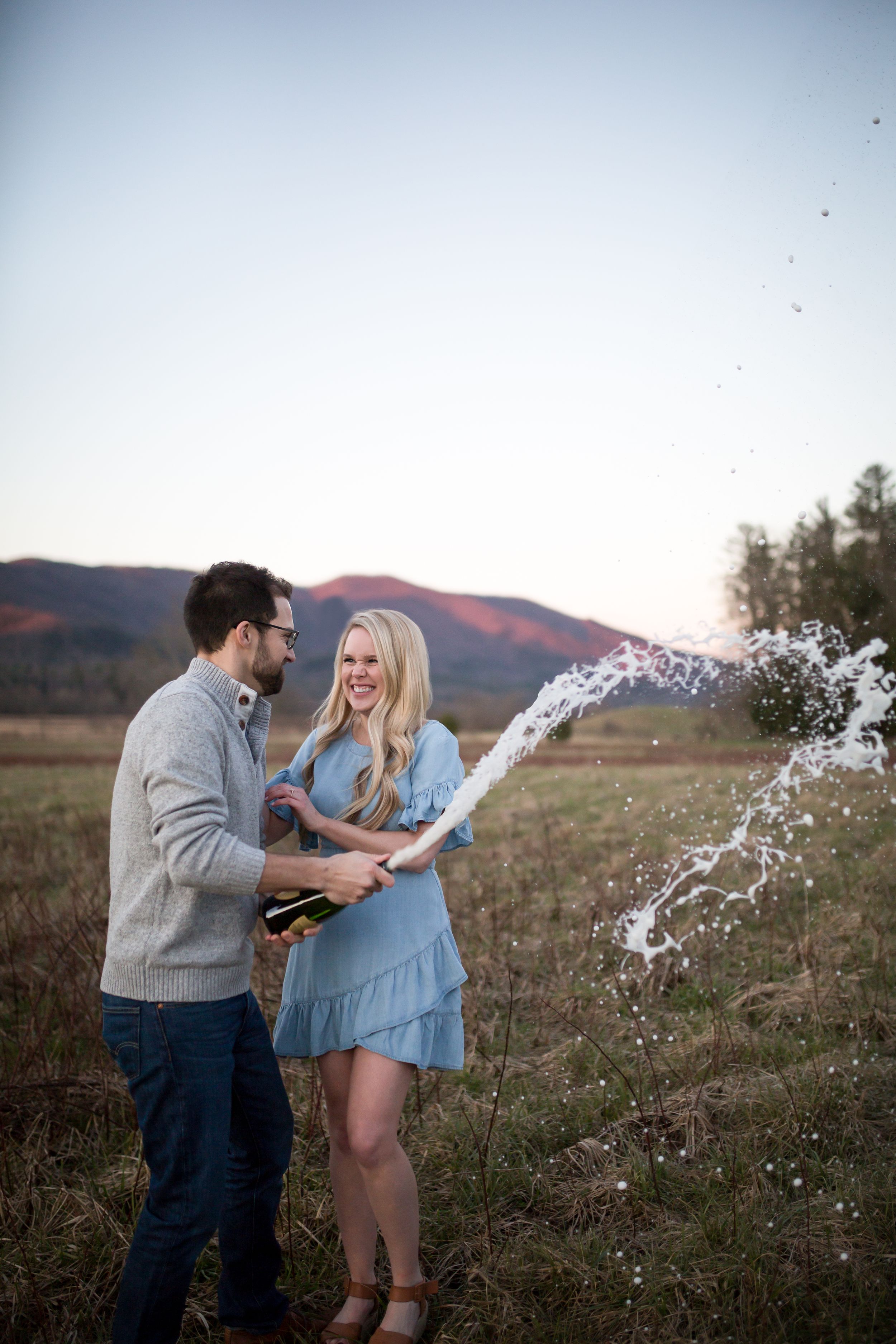 A couple celebrates with champagne spray in a field at sunset.