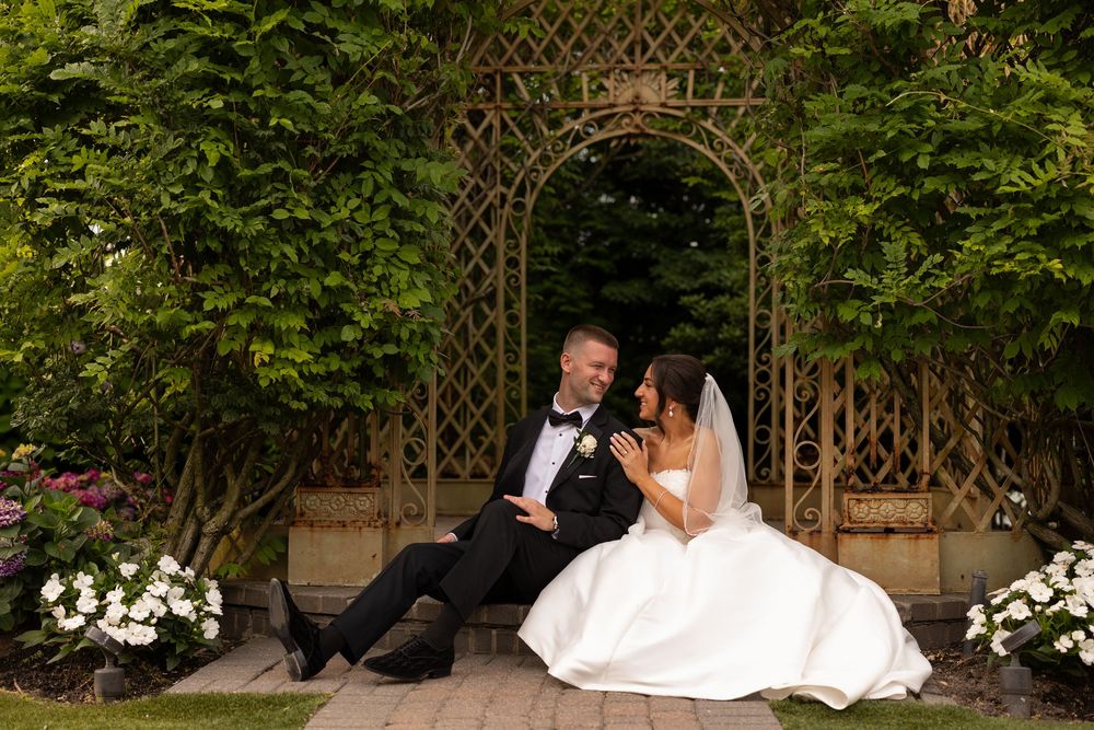 Two people embrace under a garden archway adorned with lush greenery and flowers on their wedding day.