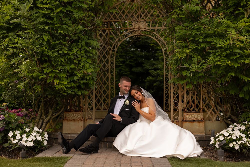 Two people in wedding attire sit together under a wooden garden arch surrounded by greenery and white flowers.