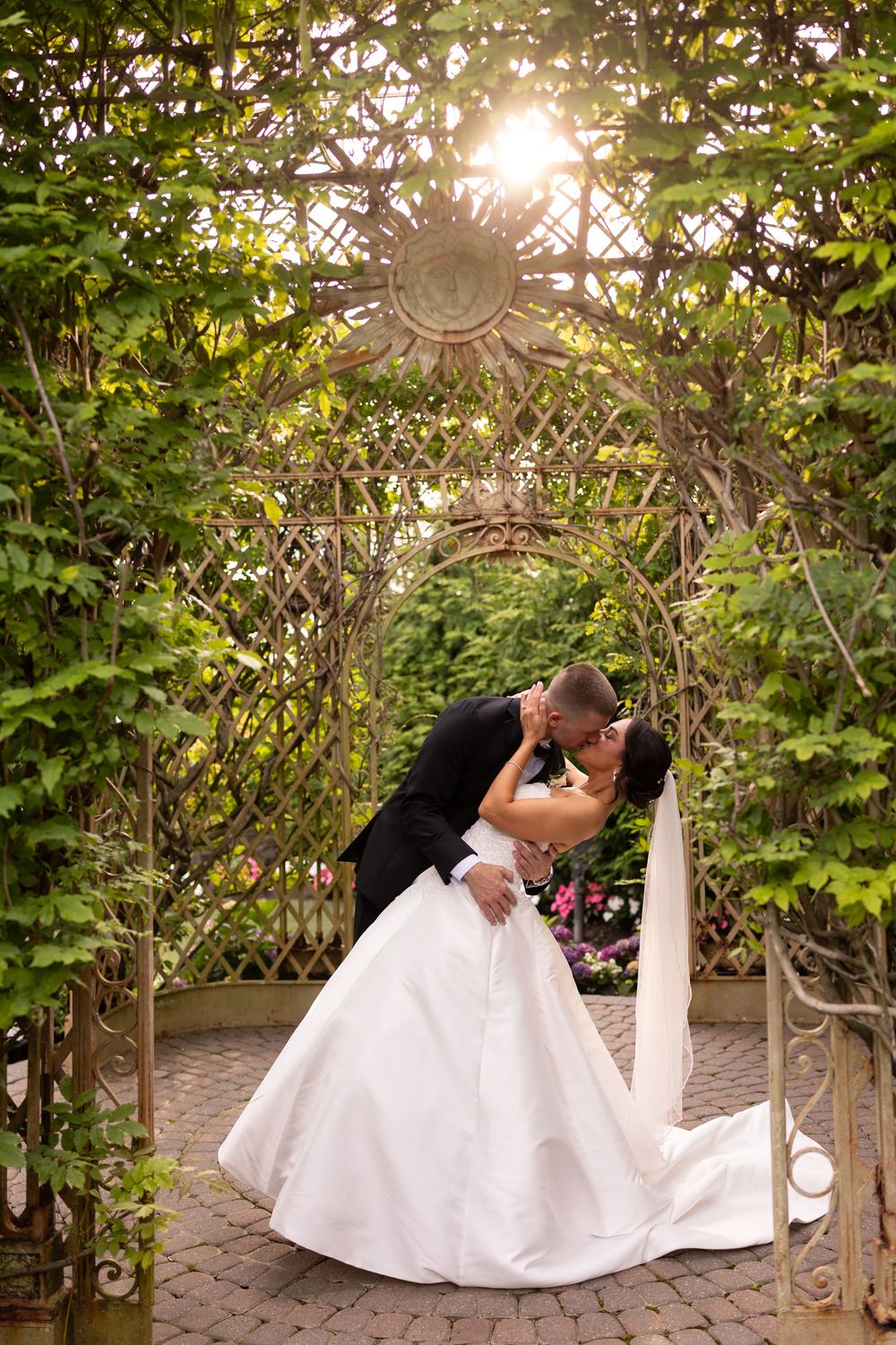 A romantic wedding couple dips under a sunlit garden archway decorated with climbing vines and floral details.