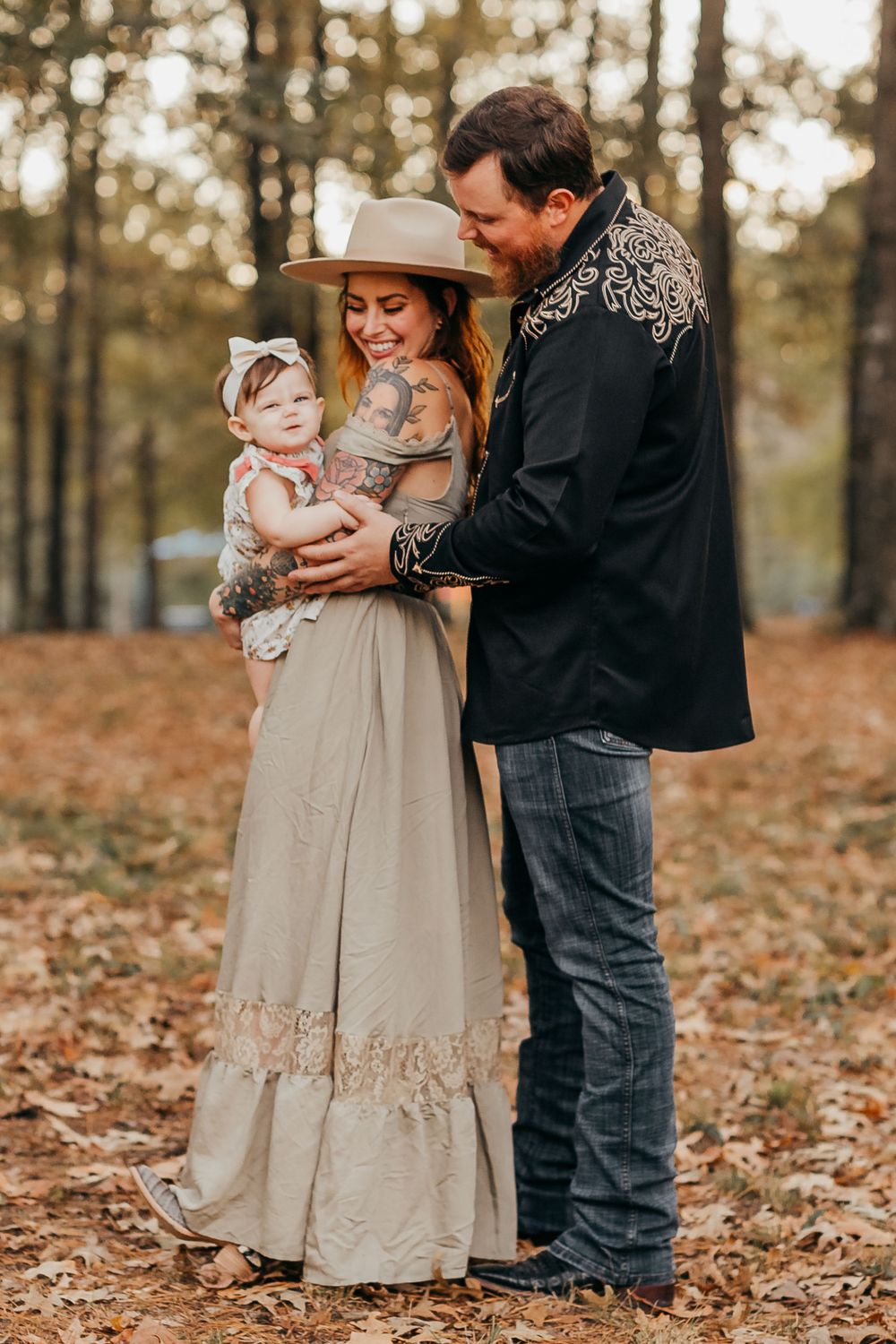 A family dressed in rustic clothing embraces while standing among fallen autumn leaves in a wooded setting.