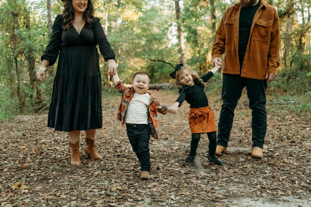 A family holds hands while walking together on a leaf-covered path through an autumn forest.