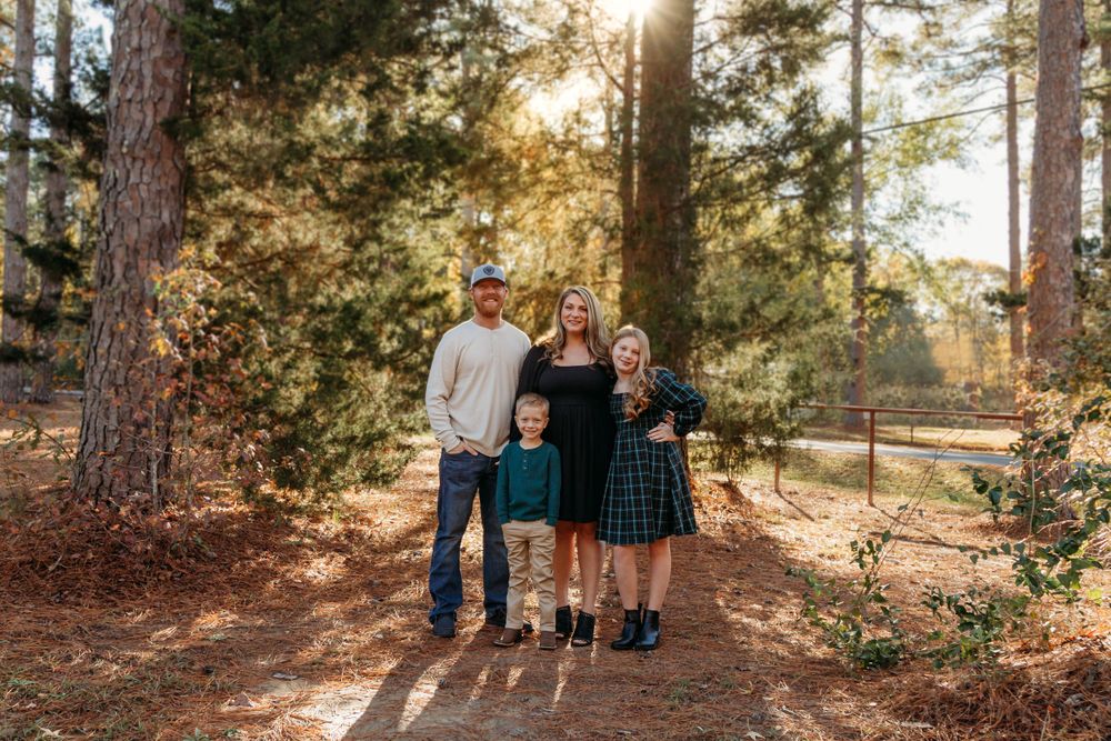 A family poses together on a sunlit forest path with sunbeams filtering through pine trees in the background.