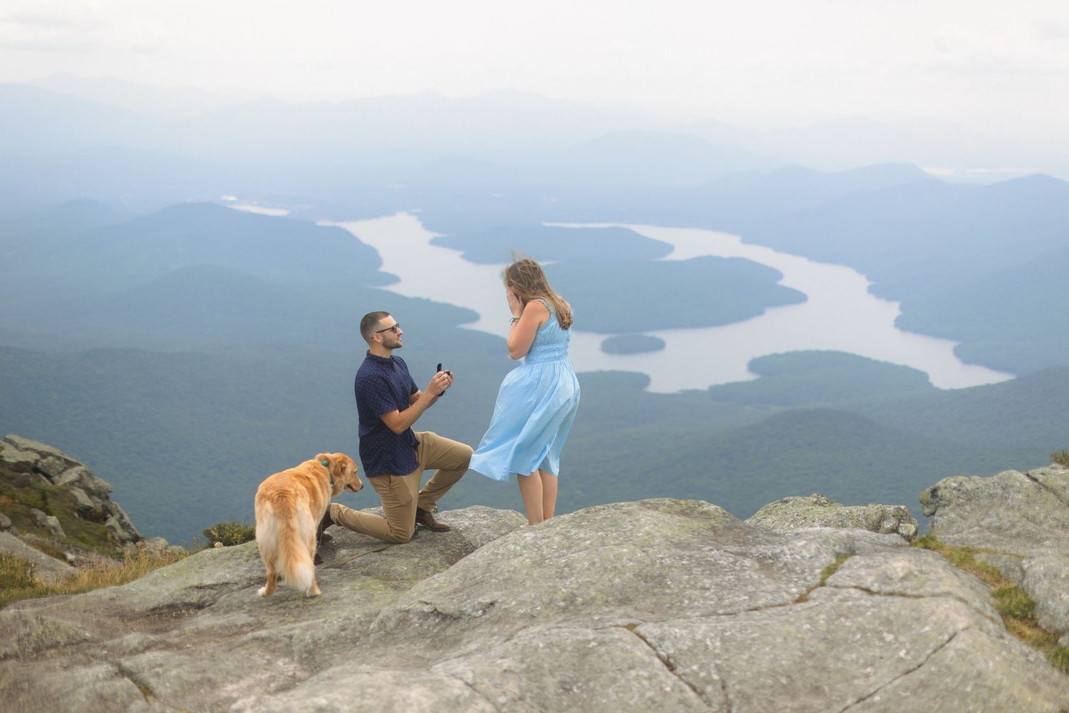 A winter proposal moment on snowy ground with pine trees and a frozen lake in the background.