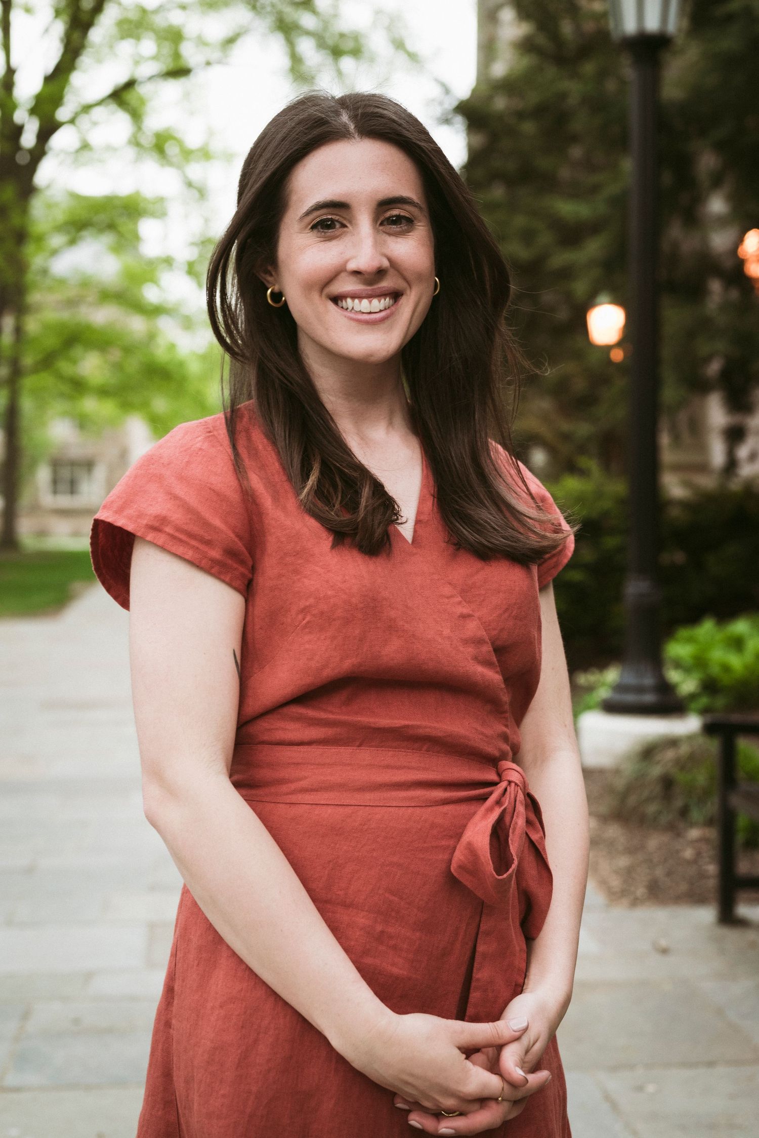 Smiling woman in a rust-colored wrap dress poses on a tree-lined campus walkway.