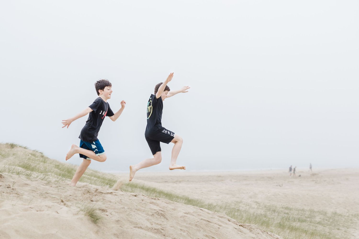 Two people jumping joyfully on sand dunes against overcast sky.