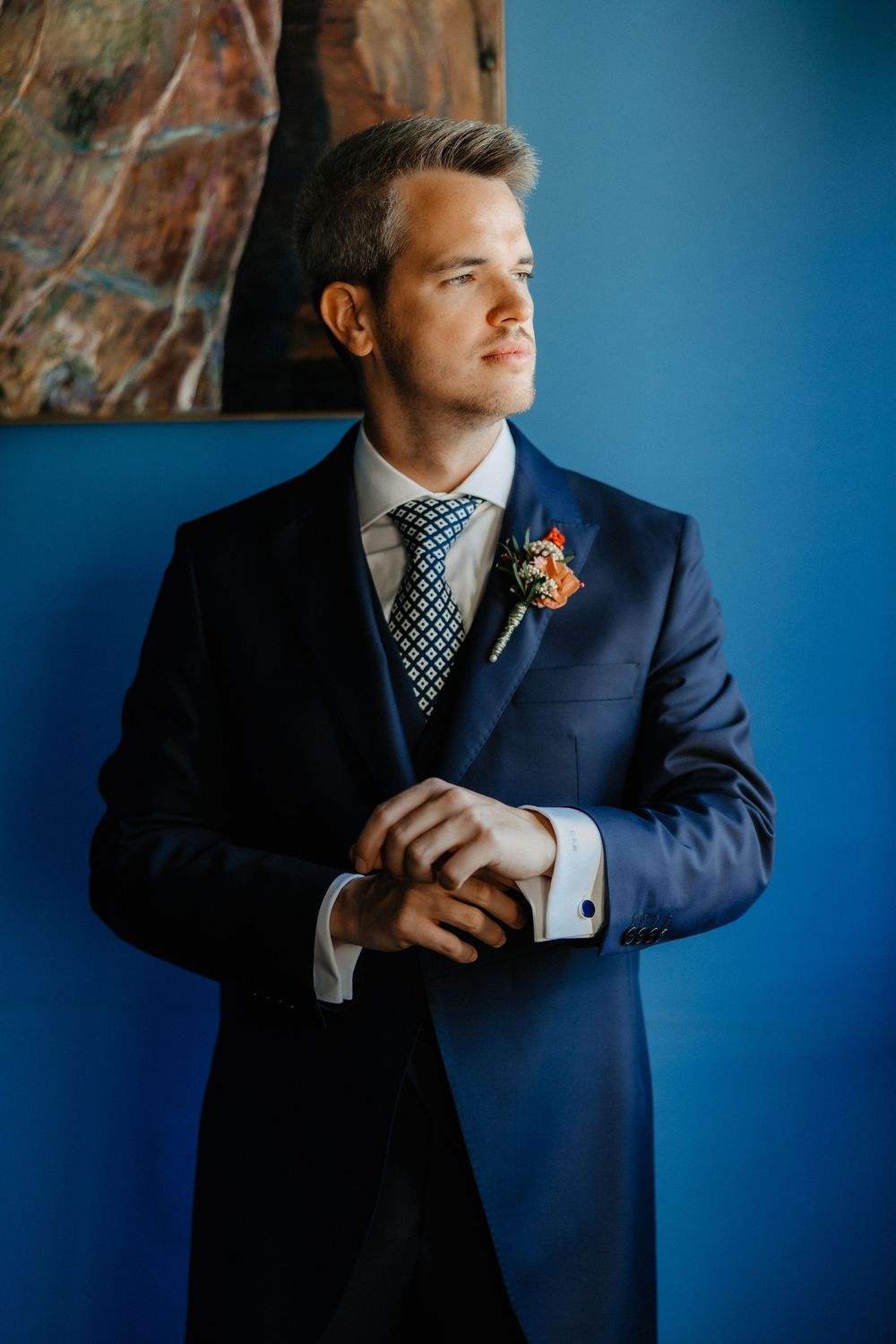 Groom in navy blue suit and polka dot tie poses against blue wall in series of formal wedding portraits.
