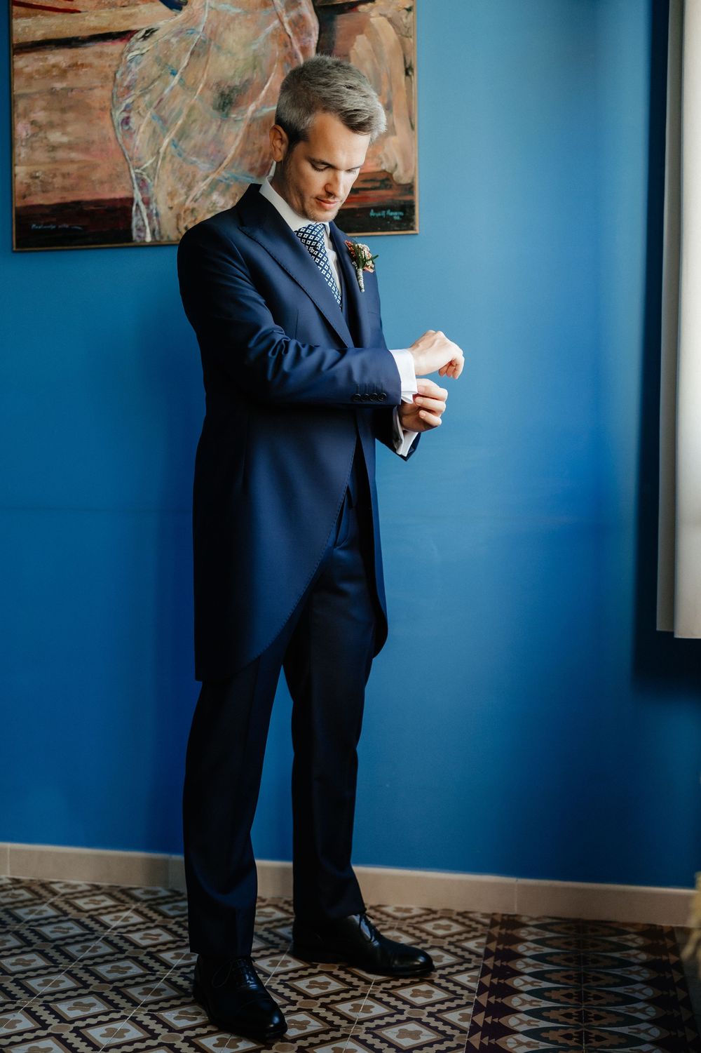 Person in navy suit checks their watch while standing against vibrant blue wall with patterned floor.